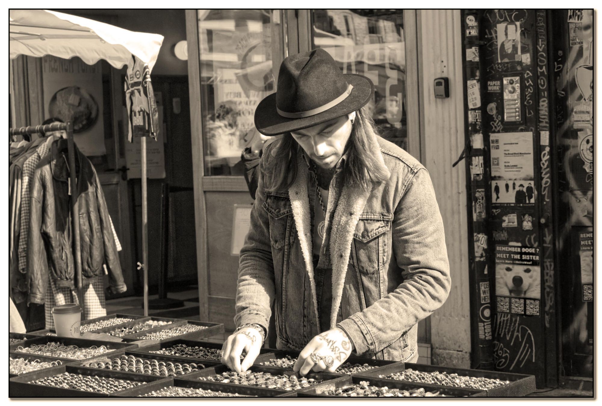 A street vendor stands behind a display table filled with various pieces of jewellery. The vendor, wearing a wide-brimmed hat and a denim jacket, is arranging or examining the jewellery items. In the background, posters and flyers are visible on a wall, along with hanging garments, suggesting an outdoor market or fair.  The photograph is in sepia tones, giving it a vintage feel.