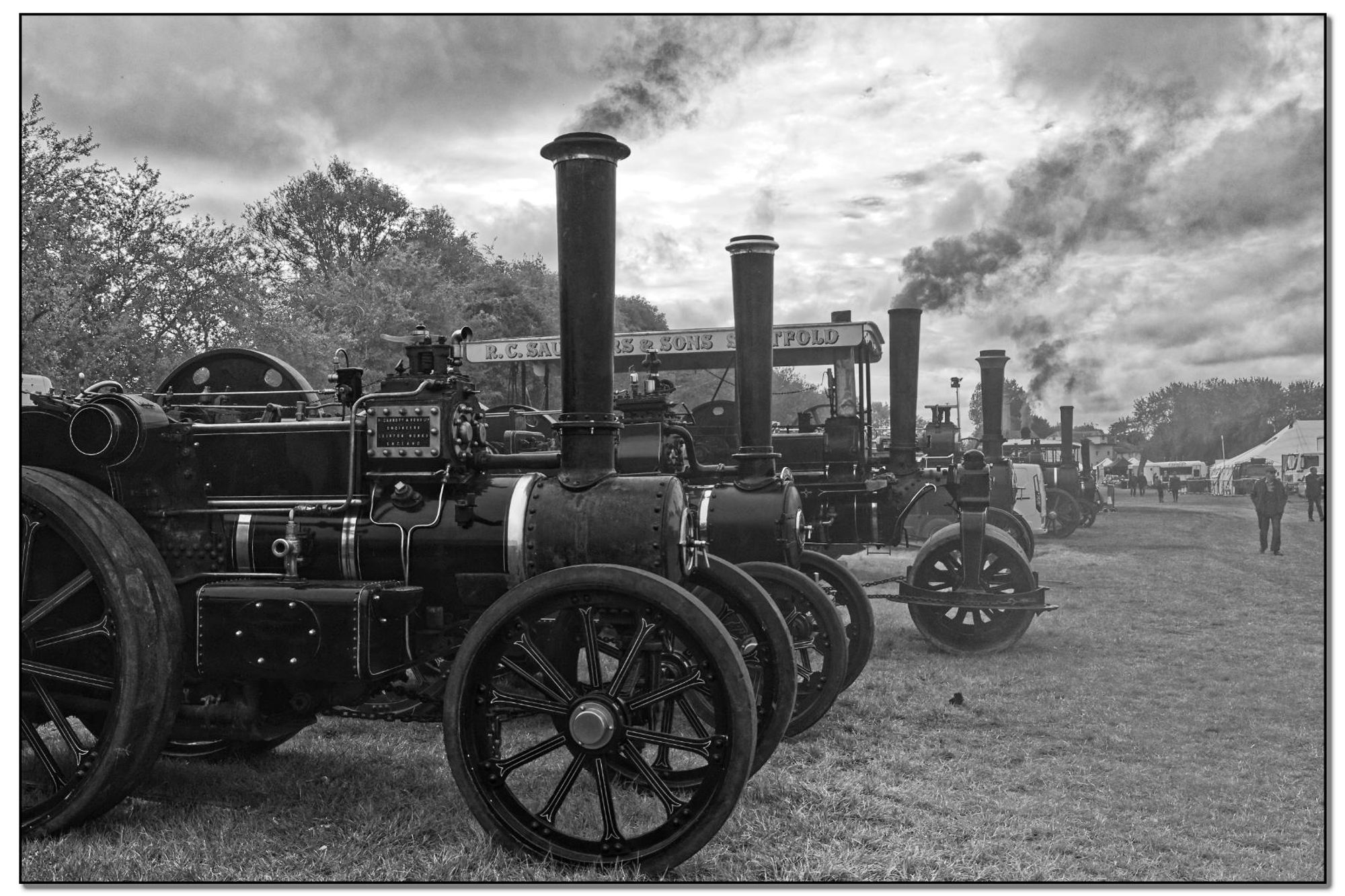 Black and white photo of a line of vintage steam traction engines lined up at a fair or event. Smoke billows from their chimneys. The engines have large wheels and are adorned with intricate details.