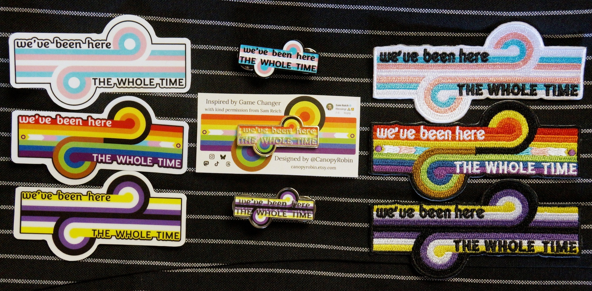 Stickers, enamel pins, and embroidered patches in trans pride, intersex inclusive progress pride, and non-binary pride colours. The shape of the design is the loop-de-loop emblem of the Dropout show Game Changer, and each design has the words "we've been here the whole time" written on it