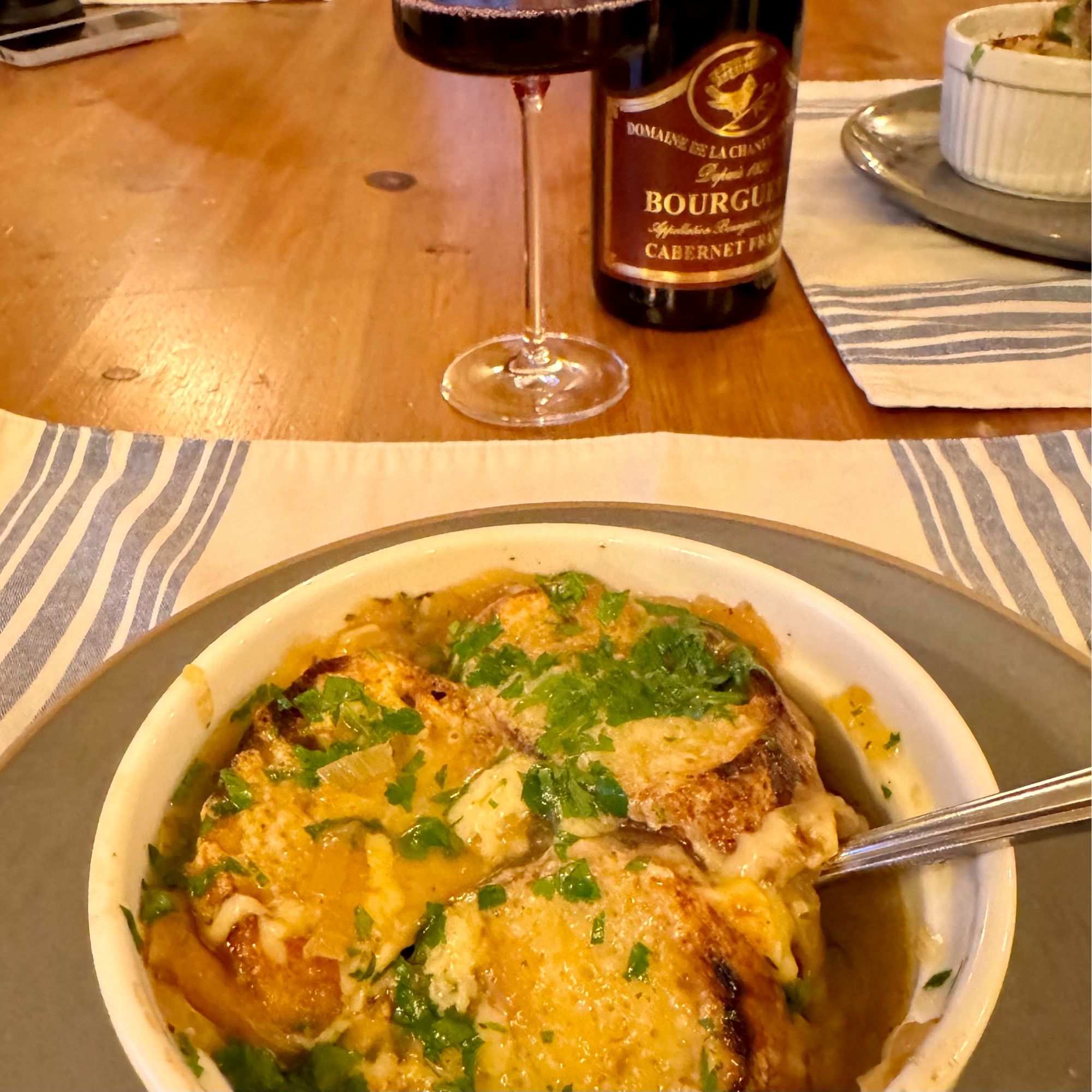 A bowl of French onion soup in the foreground and a glass of red wine next to a bottle from Bourgueil.