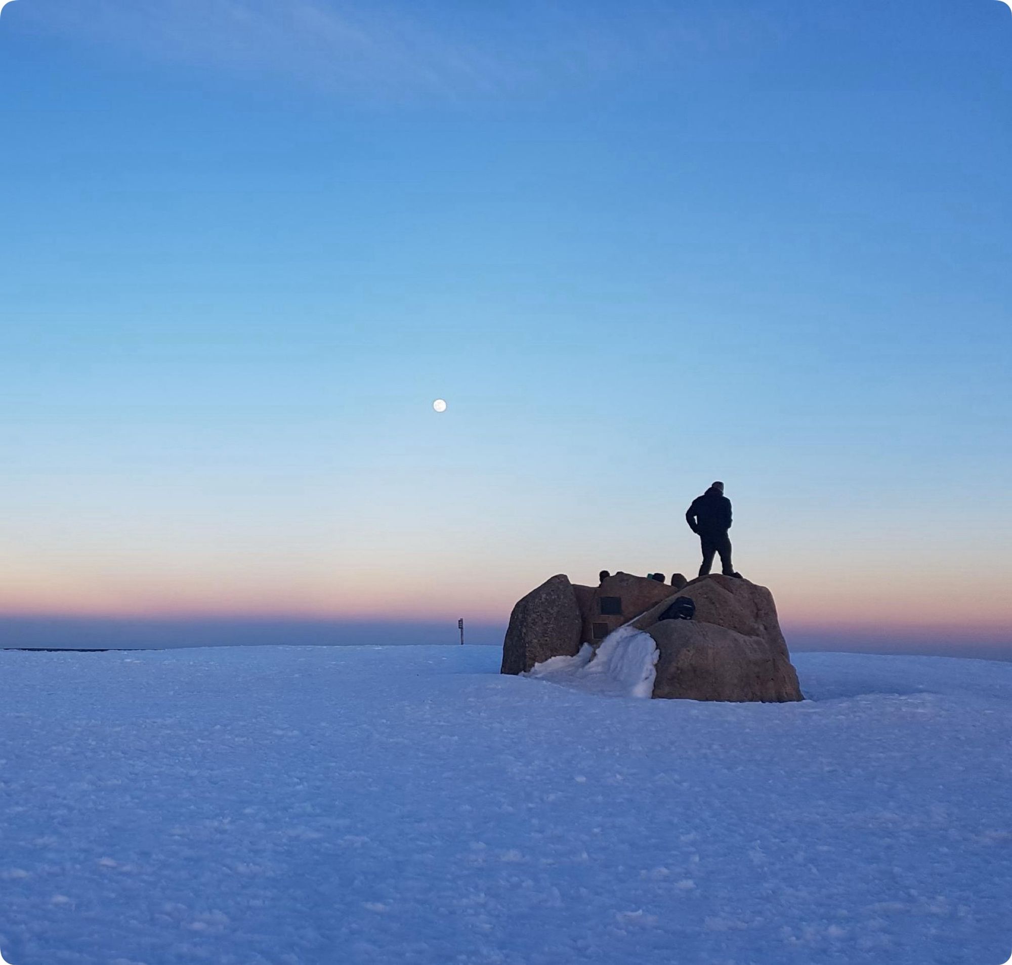 Blick über den Brockengipfel in Richtung Osten. Eine Person steht auf dem Brockenfelsen, im Hintergrund geht der Vollmond auf.