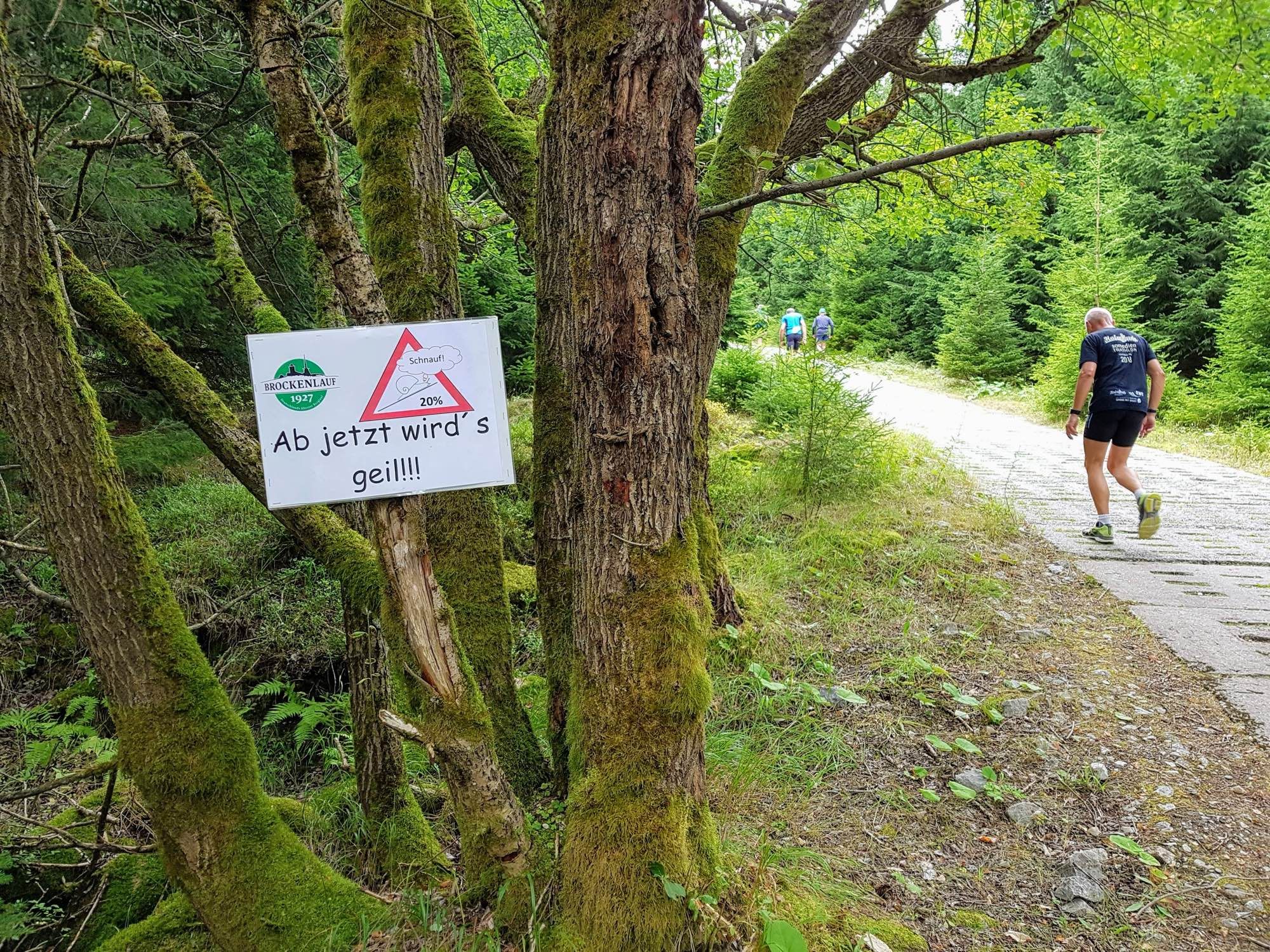 Links am Baum ein Schild mit Aufschrift „20% ab jetzt wird's geil!“. Rechts kämpfen sich Läufer den Weg rauf.