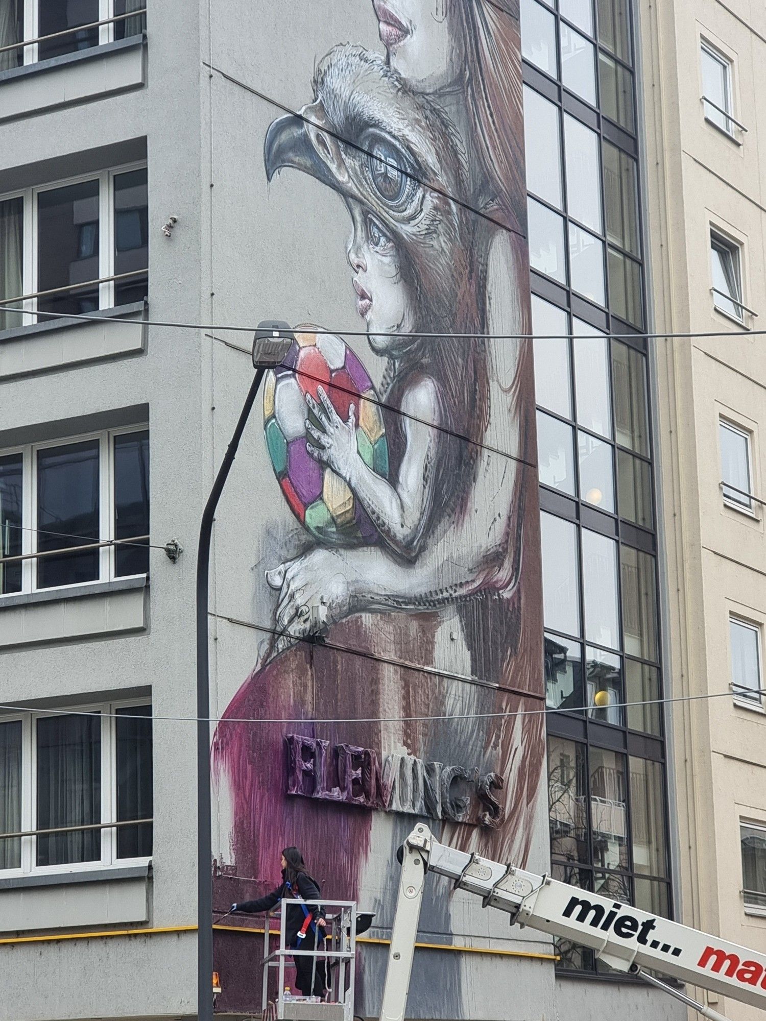 Man sieht ein riesiges Streetart Gemälde in der Frankfurter Innenstadt. Auf einem kleinen Kran steht die Künstlerin und bemalt einen Bereich unten links.