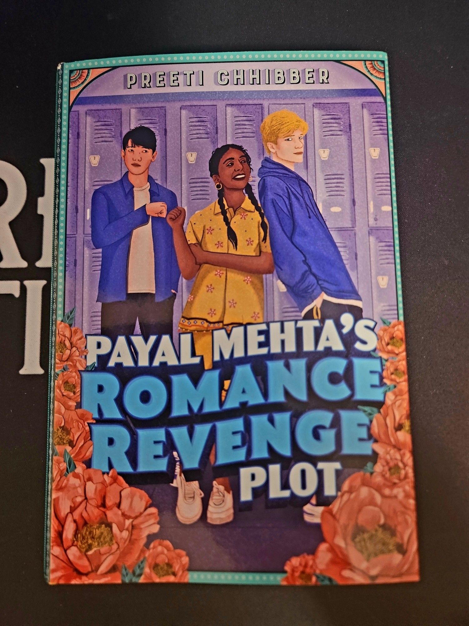 The book, Payal Mehta's Romance Revenge Plot by Preeti Chhibber, is shown on a black background.  The protagonist is shown leaning against a blonde-haired boy while fist-bumping a dark-haired boy on her other side.  There are purple lockers in the background and flowers surrounding the title in the foreground.