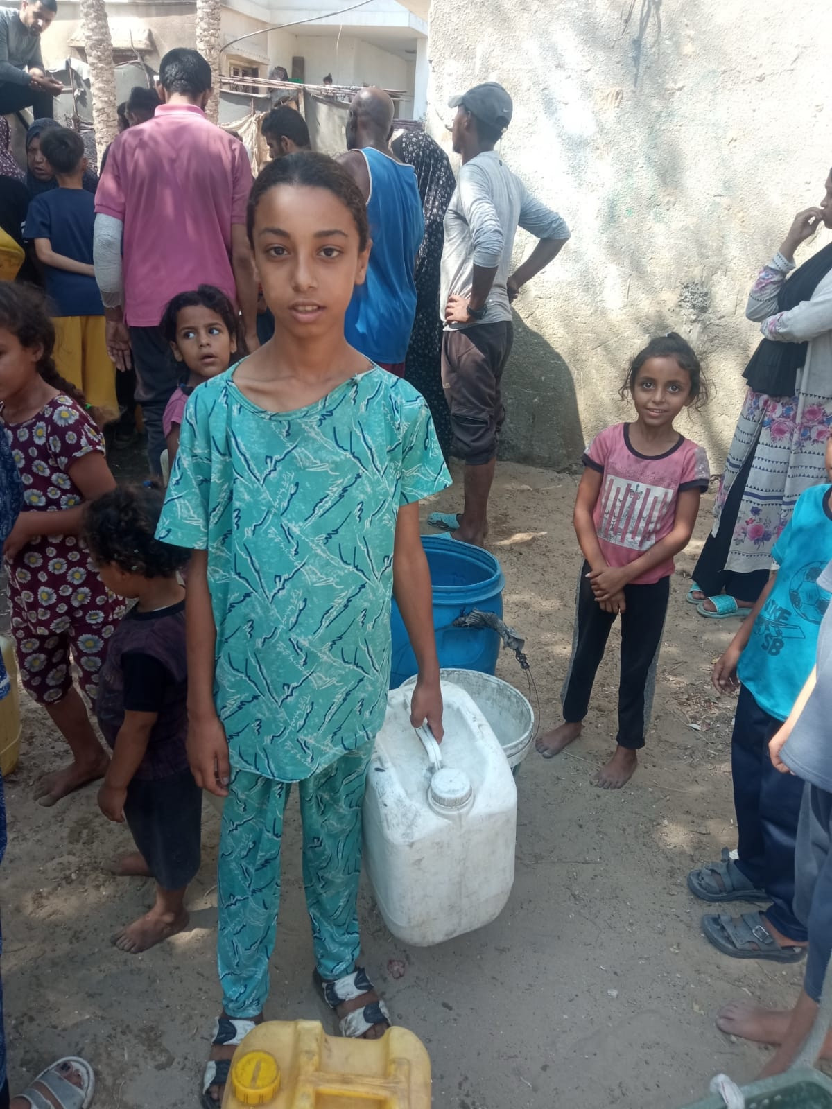 A photo of a palestinian girl, very skinny.
She holds a water canister and looks seriously into the camera. 
She is standing in a queue.
Her name is Retaj, she is Mohammed‘s sister.
The photo is taken while displacement and famine in genocide.
