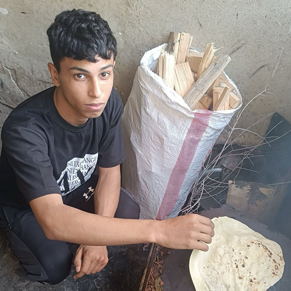 A photo of a young Palestinian man squatting in front of a cooker. Firewood collected behind him. He is skinny. The photo was taken while displacement and famine in genocide.