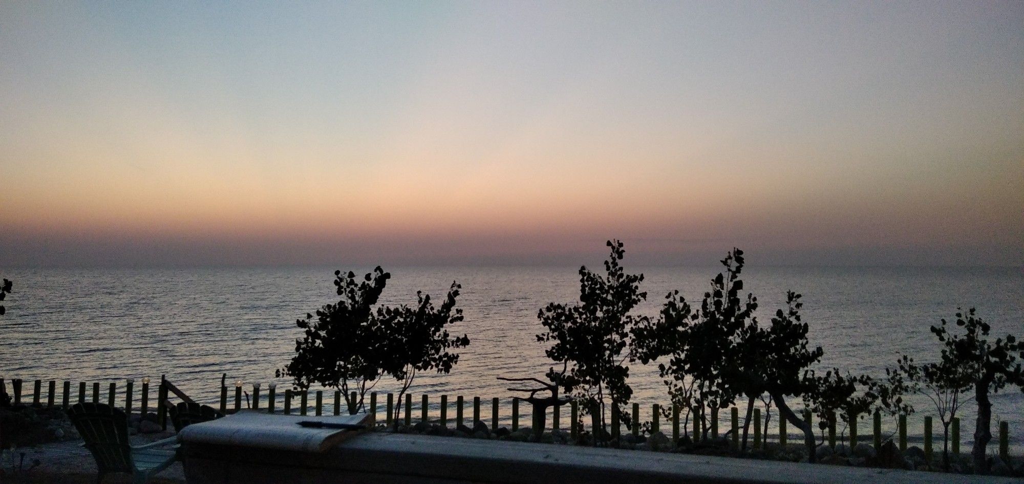 A lake at sunset with the sky and some small trees and a fence in the foreground. The sky is pale blue fading to yellow and orange at the horizon.