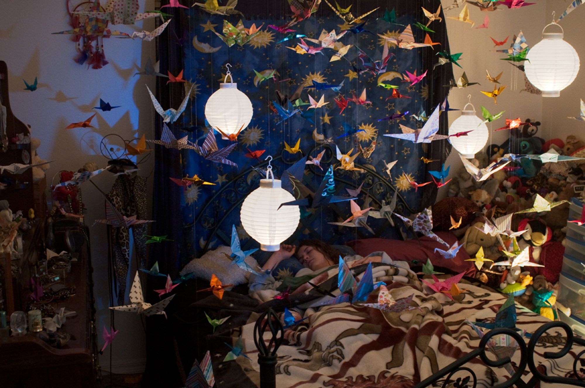 A woman lies sleeping in her bed. Above her hang an extensive assortment of paper cranes, filling the entire image, and four glowing paper lanterns.