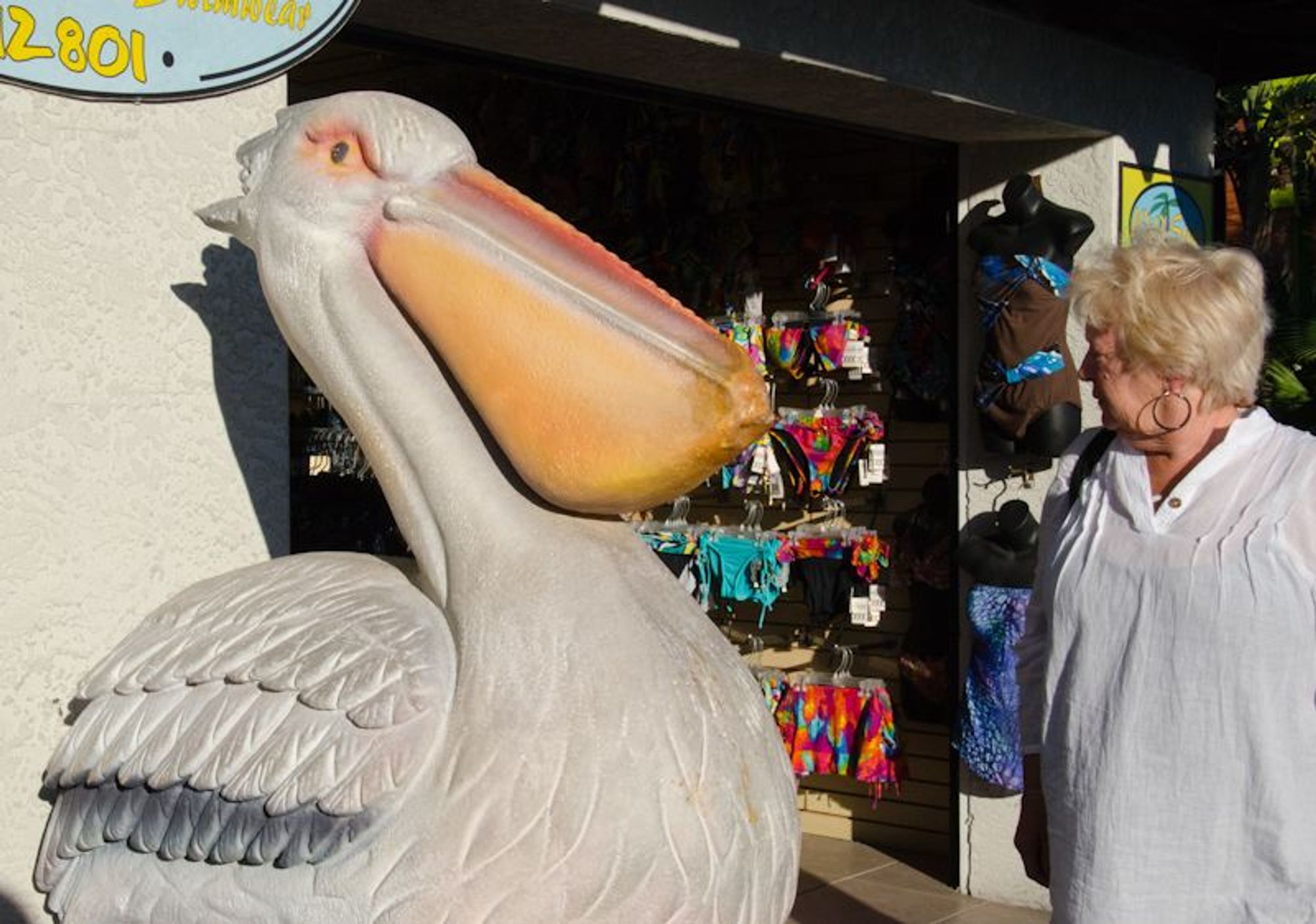 A woman in a white shirt with peachy skin and blonde hair staring down a large pelican statue, taller than she is, with white feathers and a peachy colored bill. Behind them is a store selling bathing suits.