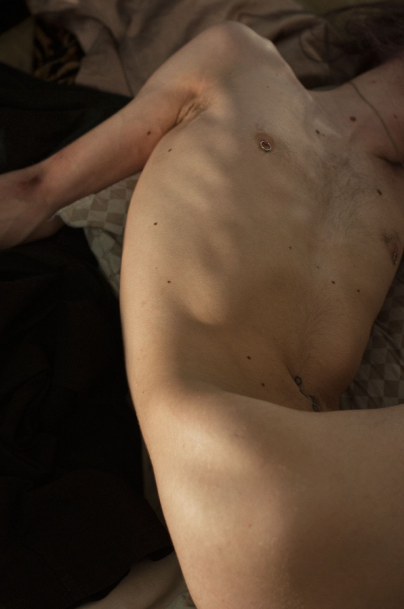 From Heroin, a series of images simulating heroin abuse. This image shows a skinny man, one arm splayed out, the inside of the elbow darkened and bruised. His face cannot be seen - his head falls out of the view of the camera. His prominent bones are visible in the falling of light across his body. His nipple is pierced.