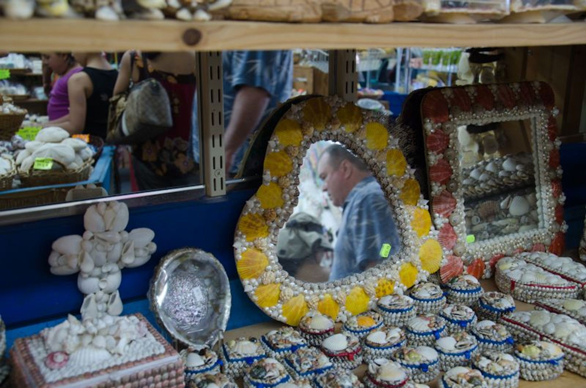 A display in a storefront selling mirrors and small jewelry boxes with shells glued to them, covering their surfaces. In the center, heart shaped mirror in the reflection of a man, his face in profile and framed by the upper "arm" of the heart. More mirrors reflect other customers and products as well as the centrally pictured man's arm and shirt.