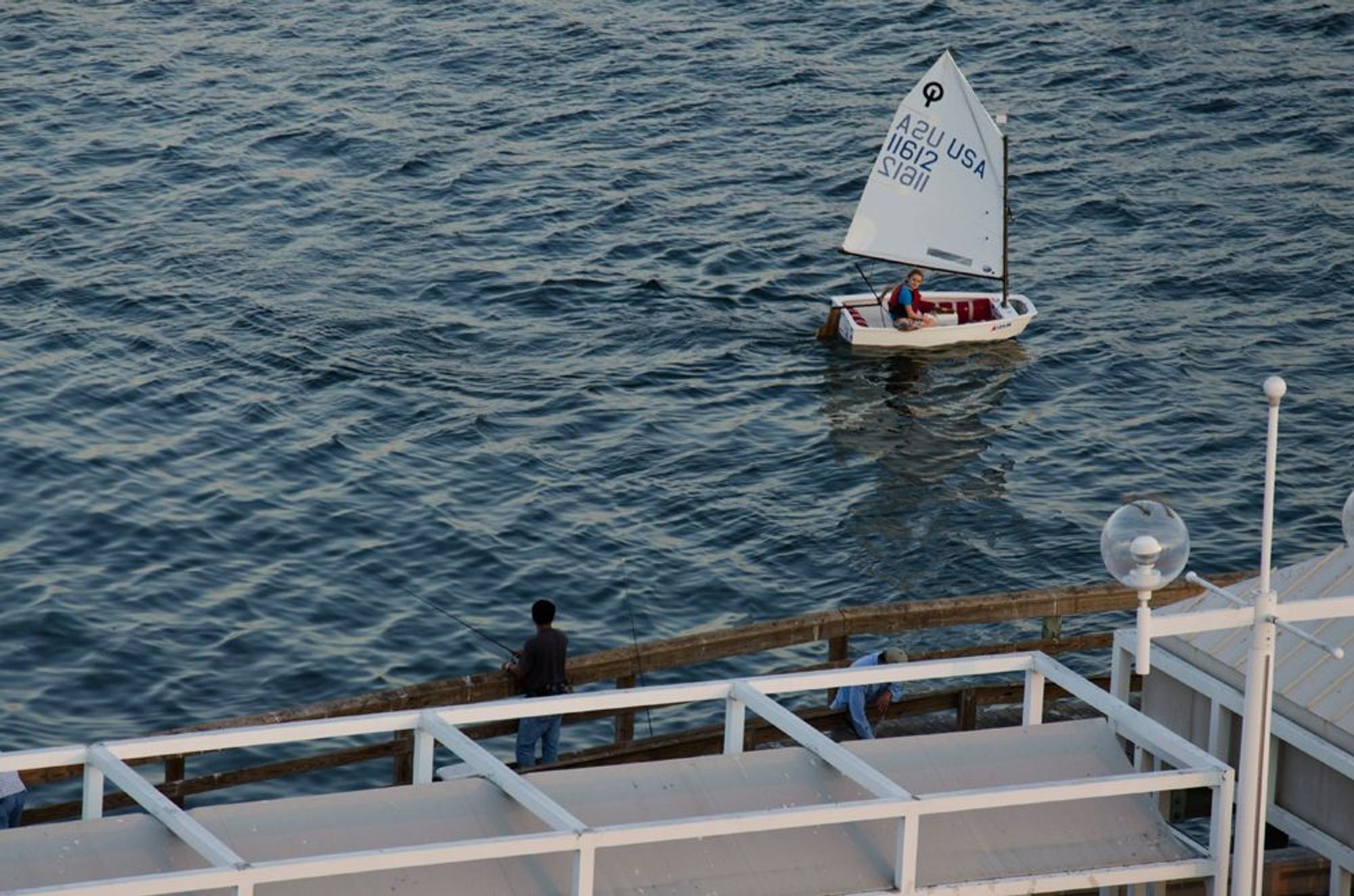 A picture taken from the St. Petersburg Pier before it was renovated. A man is fishing from a dock while a woman sails past in a small single passenger boat. Her head is turned back towards the pier, perhaps even to the man fishing.