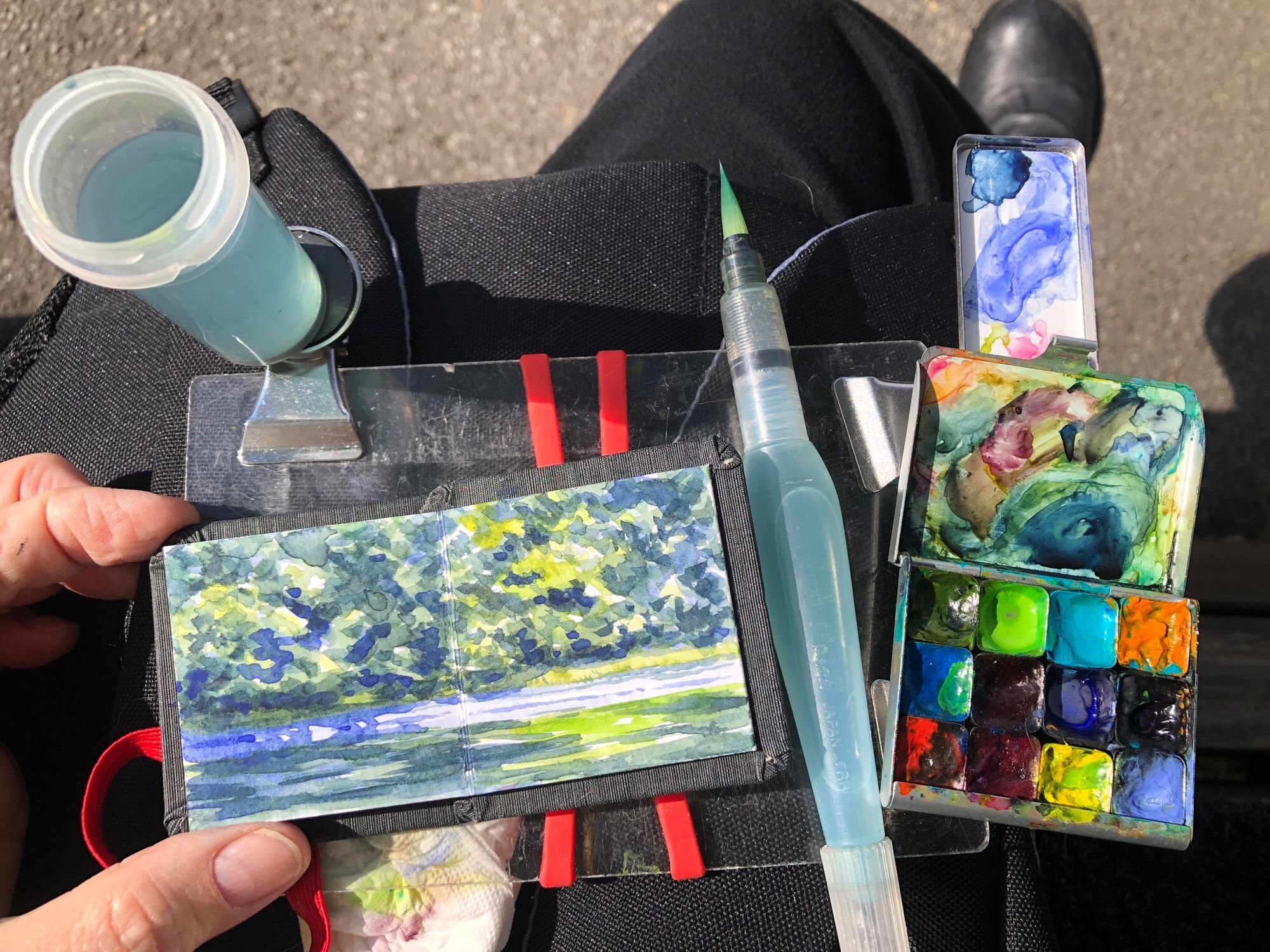 My tiny sketchbook is on my lap along with my paint, palette, watercolor, brush, and water container. The sketchbook is open to the page that I have just painted of the edge of the pond at Parc Lafontaine.