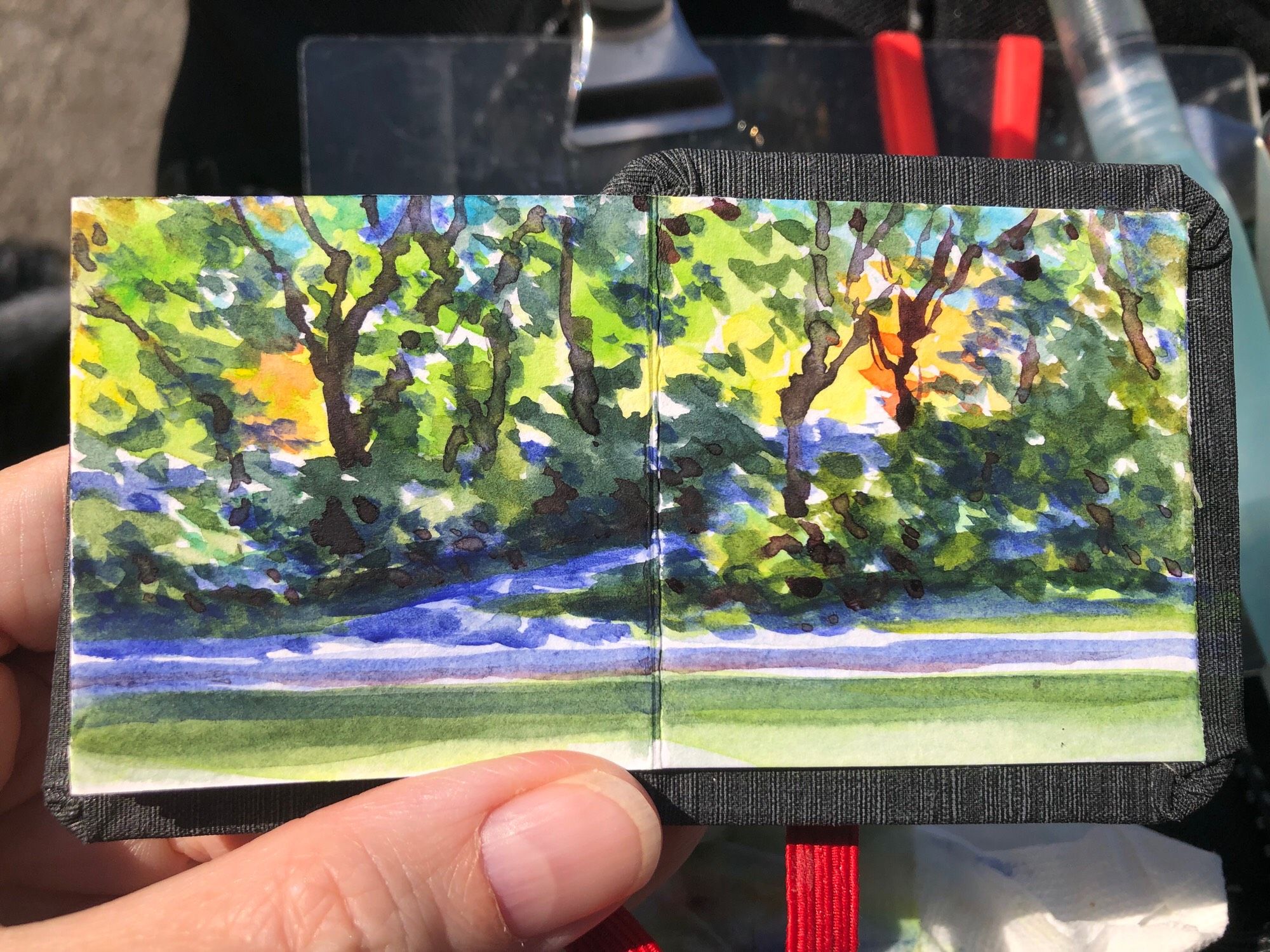 Small, watercolor, painting, approximately 2” x 4”, in my tiny sketchbook. This is a painting done on site at Parc La Fontaine here in Montreal, and it depicts the view across the pond. There are some trees and bushes and a path leading up from the edge of the pond. There is sunlight coming through the tops of the trees and shade dappled along the bushes and the path. It is an autumn scene, and there are warm and cool colours coming through.