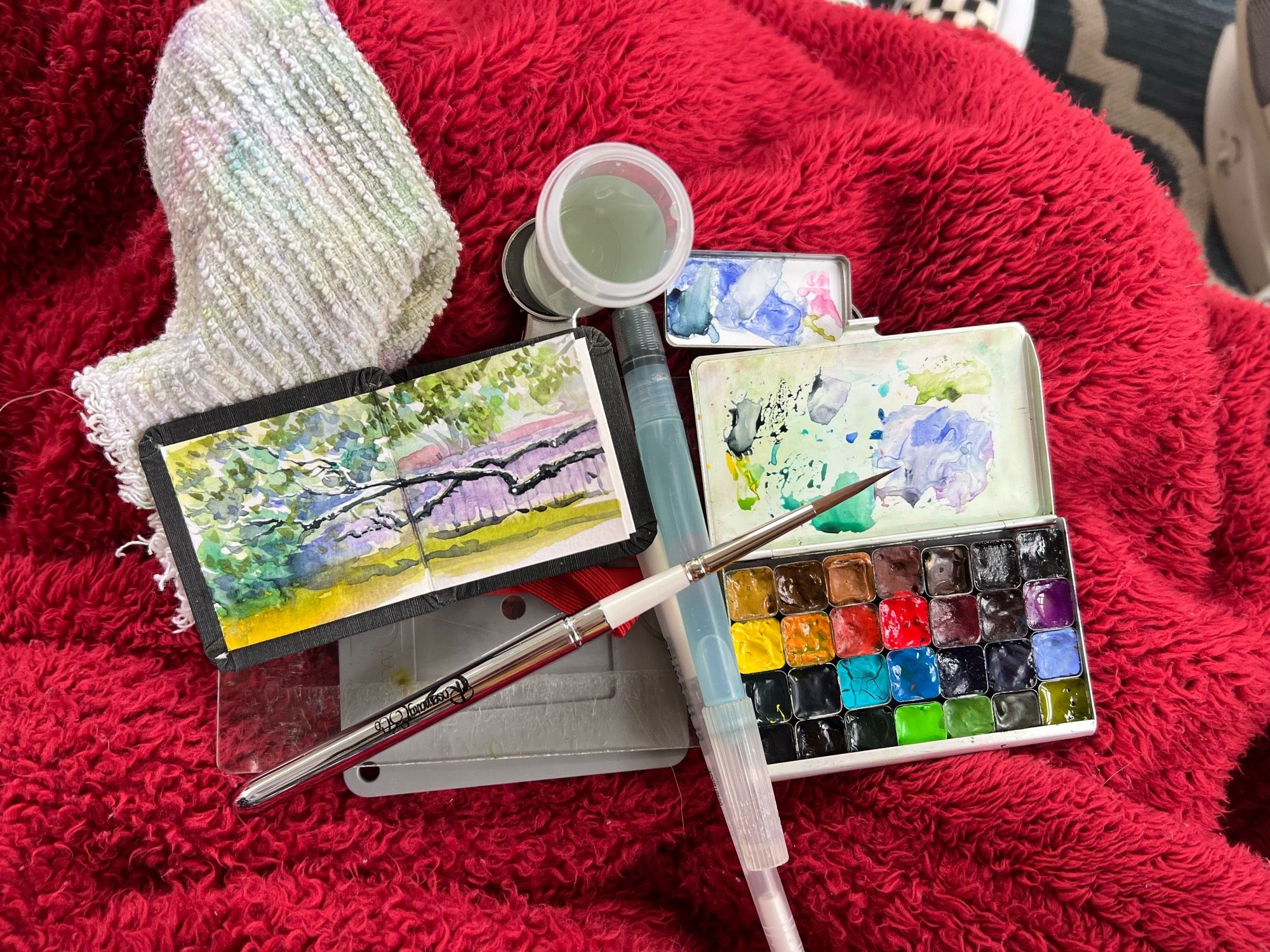 My tiny, watercolour sketchbook it’s on my lap with my paint palette, and a few watercolour brushes. Painting is of some branches of a large tree, and they are horizontal.