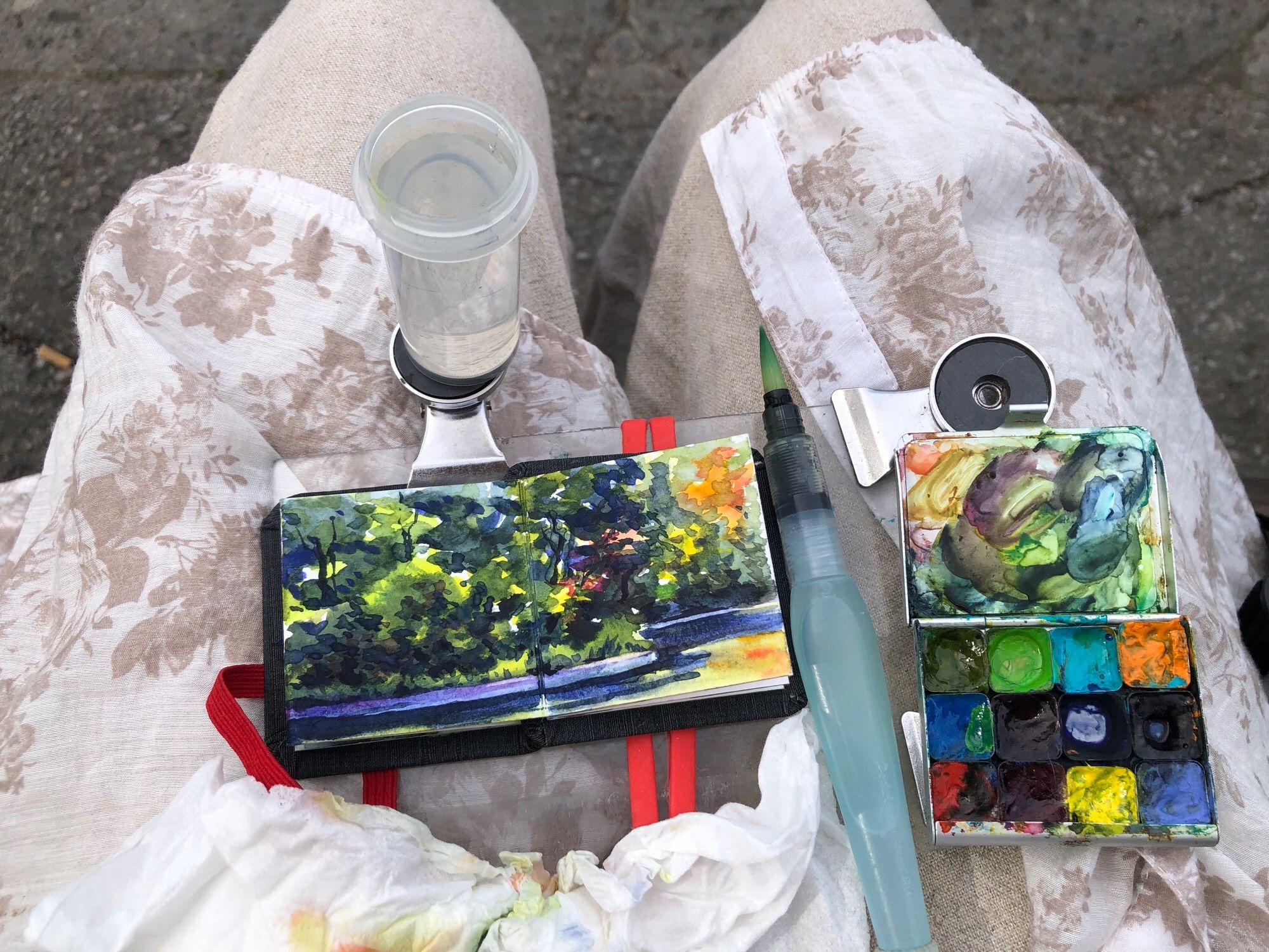 My watercolour sketchbook is in my lap, along with my paint, palette, brush, and water container. The sketchbook is open to the page where I have just painted a view of Parc Lafontaine pond. It is an autumnal scene with foliage and trees, reflecting in the water.