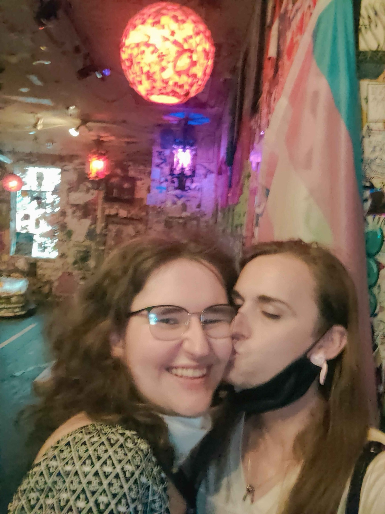 Blurry shot of Colie kissing Rayna on the cheek in front of a trans flag hanging in a bar. Rayna is looking over its shoulder and smiling at the camera