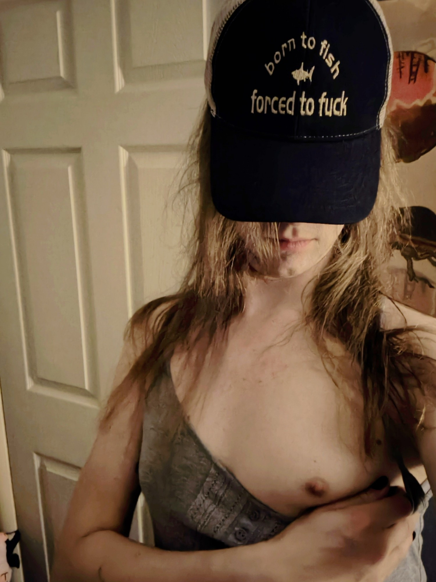 Colie standing in front of a door and flashing a tiddy. She's wearing a trucker hat that says "born to fish Forced to fuck" with a little fish in the middle of it