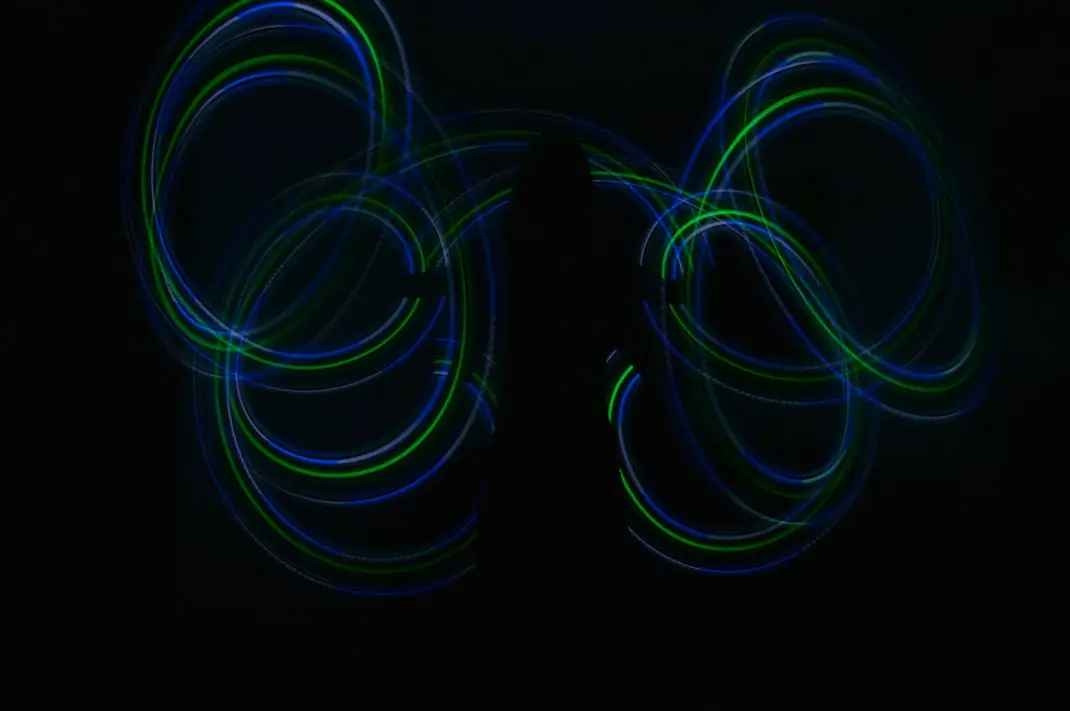 Long exposure photo of Colie dancing with poi. She is cloaked in darkness and there are tracers of green and blue lights forming circles around her outstretched arms and back 