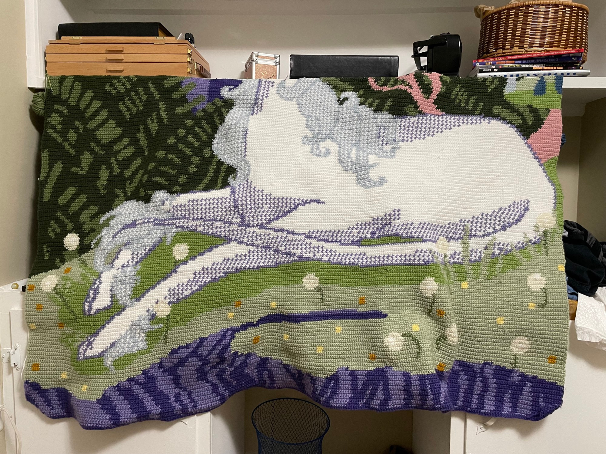 A crochet blanket in progress, in contrasting jewel tone and pastel greens, purples, blues, yellows, and pink. The headless (for now) Unicorn sits atop a mossy hill covered in dandelions in various stares of bloom and is surrounded by deep green bushes, steel purple cliffs, and mystical lilac trees. 