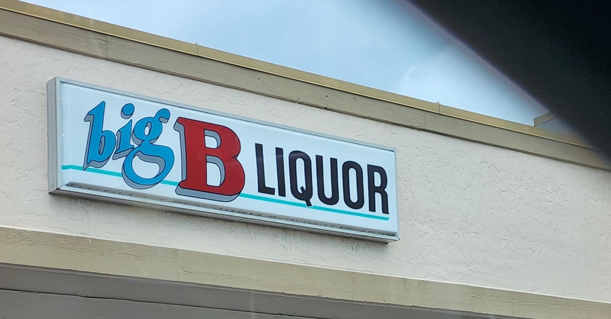 A white rectangular sign that reads "Big B Liquor" with "big" in blue script, "B" a big red Block lerter and" liquor" in thin black block font