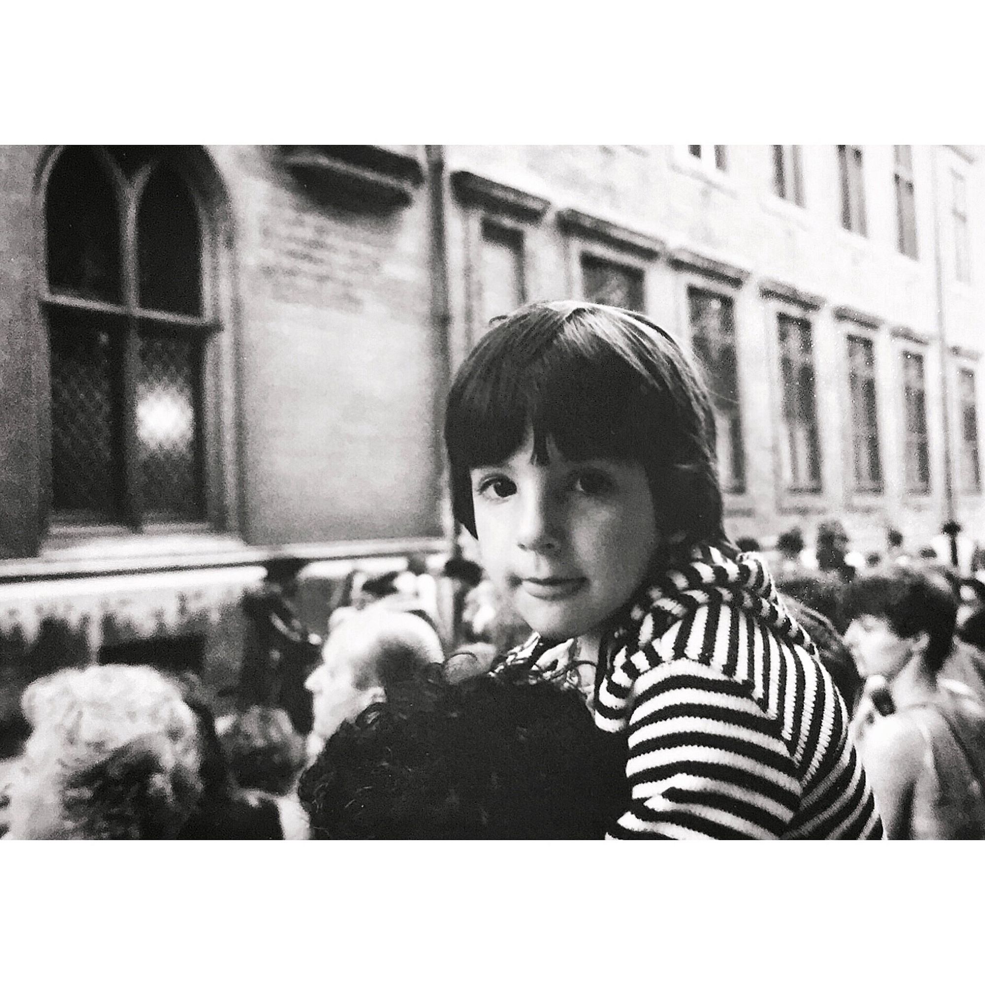 Black and white photo of a young boy sitting on his parent’s shoulders at a parade.