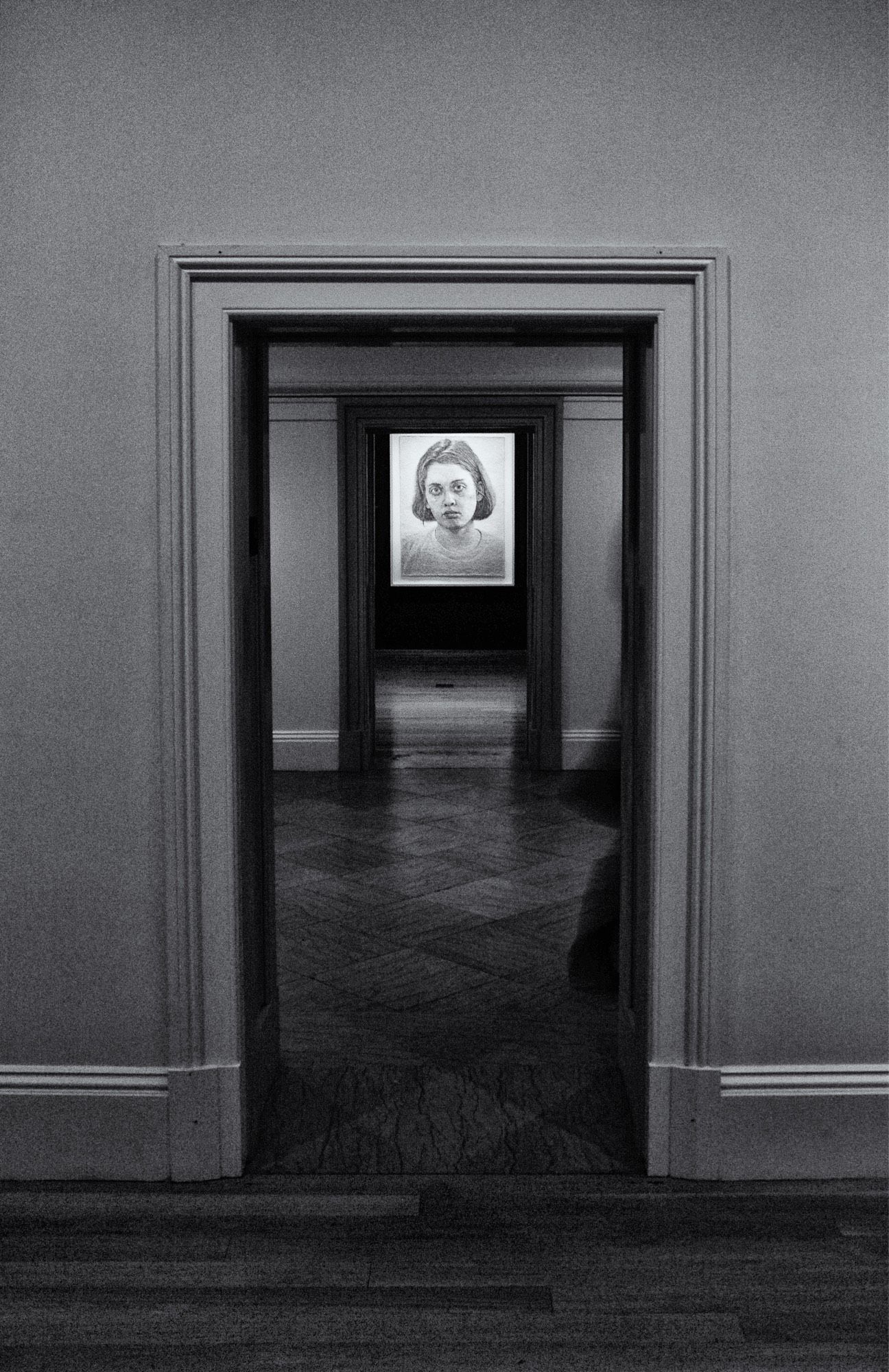 Black and white photo of the interior of an art museum. In the distance there is a lighted painting of a woman’s face.