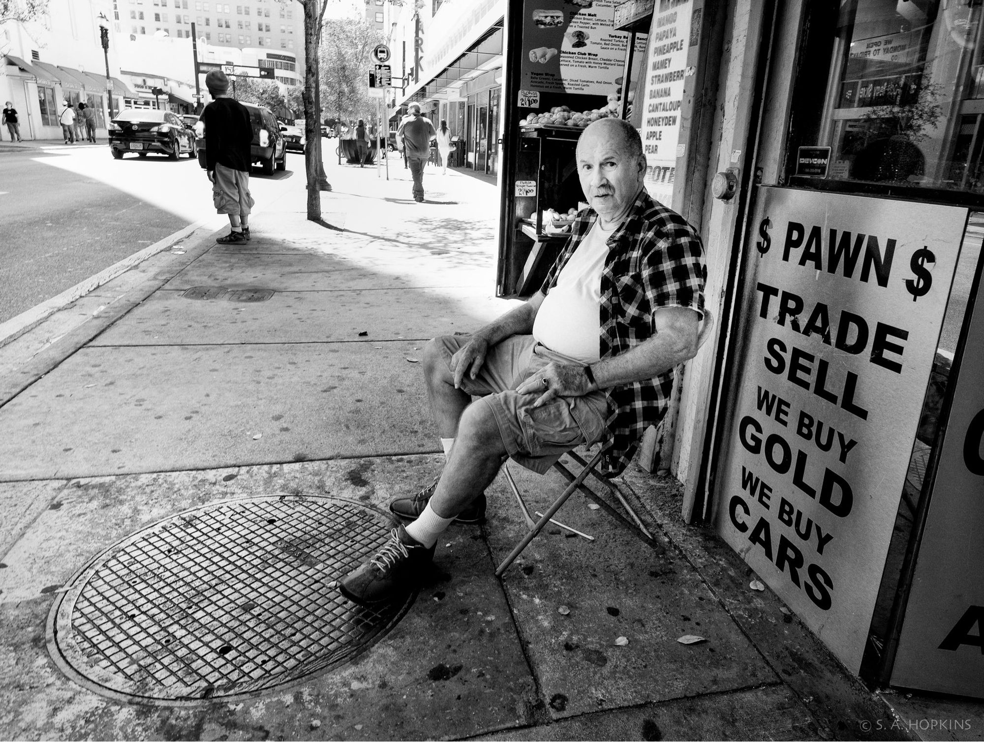 Black and white photograph showing a sidewalk scene in a downtown area. We see a bald older man seated in a chair in front of a pawn shop.