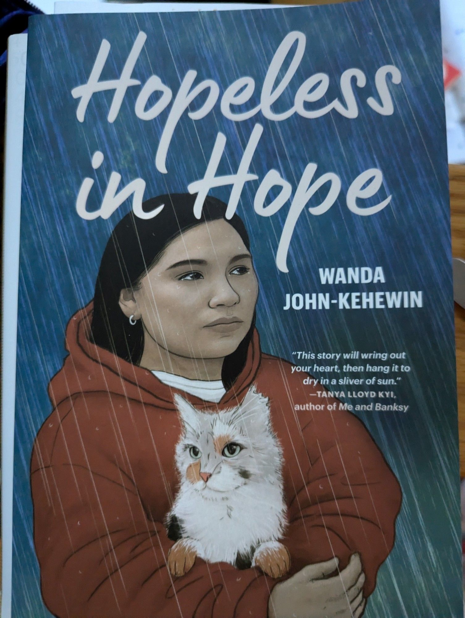 an indigenous girl with black hair and a red hoodie holding a white cat in the rain. Text reads Hopeless in Hope Wanda John- Kehewin. "This story will wring out your heart, then hang it to dry in a sliver of sun" Tanya Lloyd Kyi, author of Me and Banksy