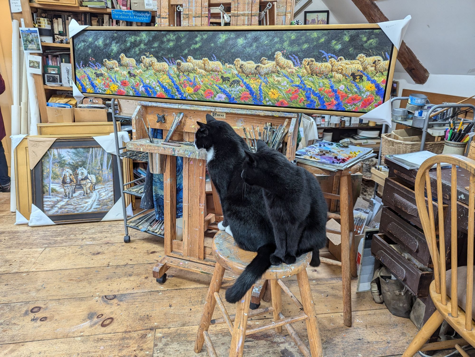 Two black cats sit on a stool in front of a horizontal painting of sheep and flowers in a field. Oil painting paraphernalia is visible in the background. Another painting of horses pulling a wagon on a snow dusted track sits on the floor by a full bookshelf.