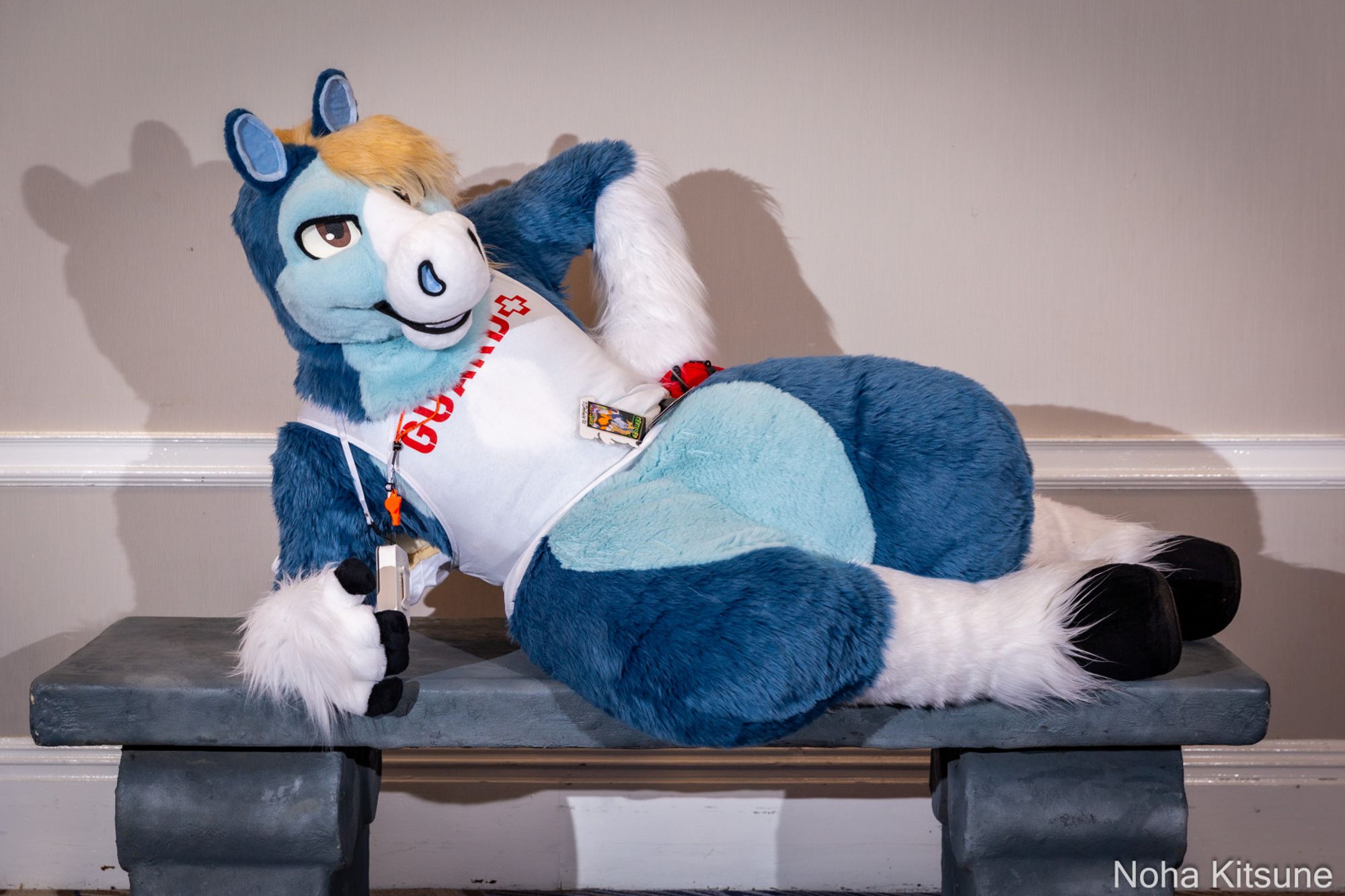 A photograph of a blue pegasus fursuiter at ConFuzzled 2024, laying on top of a grey bench facing the camera while propped up slightly with his right arm. He's wearing a white lifeguard tank top with a red whistle on a lanyard around his neck, and behind him is a plain white wall with cast shadows.