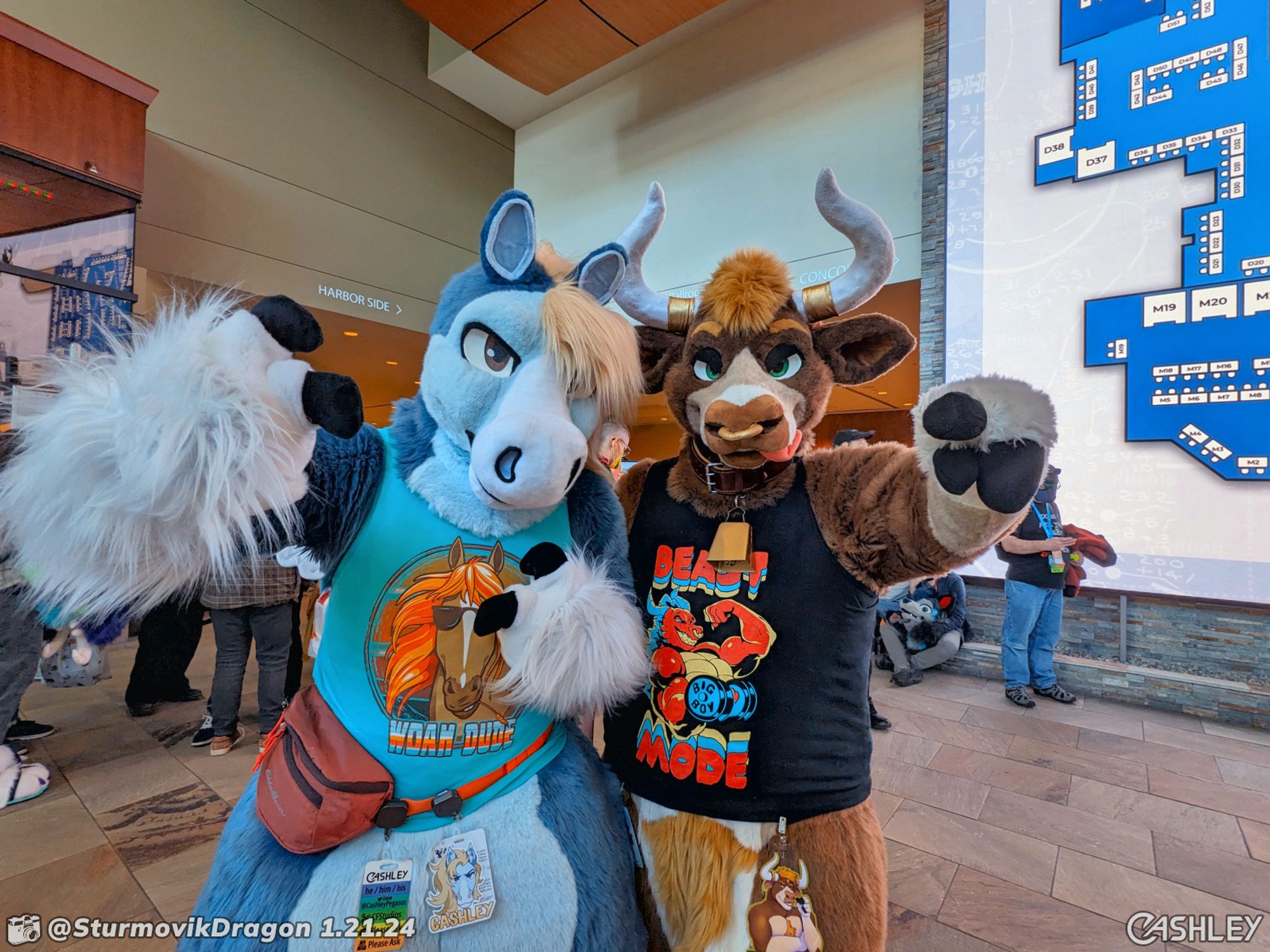 A candid photograph of two fursuiters posing together and pointing at the camera while standing in the main lobby of the Anthro New England Westin. On the left is a blue pegasus wearing a sky blue "Woah Dude" tank top, on the right is an auburn bull wearing a black "Beast Mode" tank top.