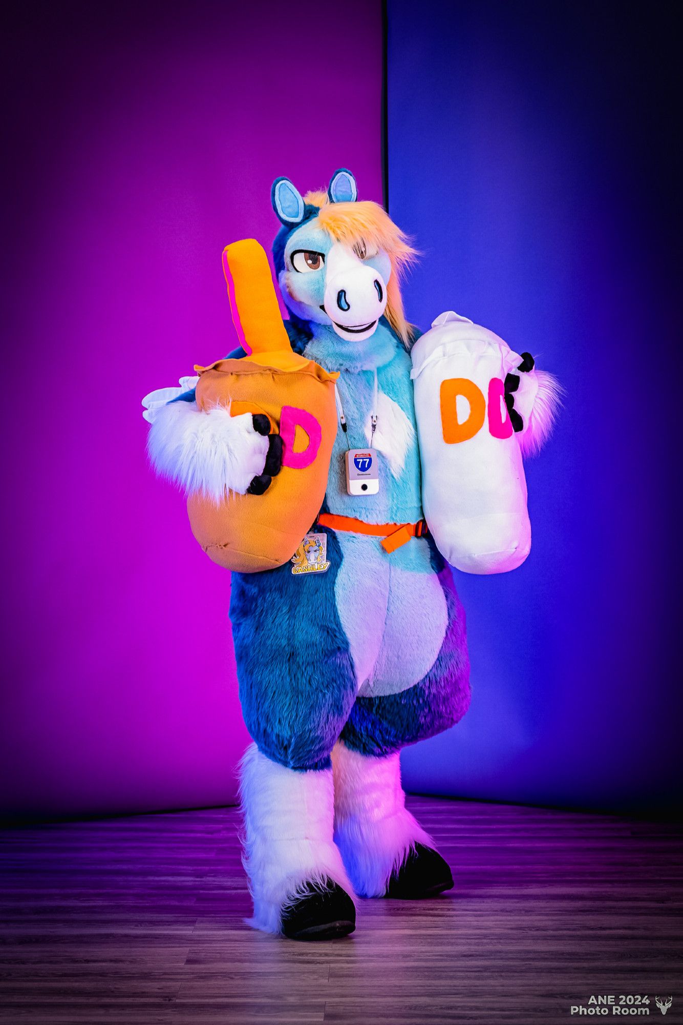 This is a photograph taken in the photo room at Anthro New England 2024, with a purple and blue color-blocked backdrop. In the center of the picture is a blue pegasus fursuiter looking at the camera while holding an oversized plush Dunkin's ice coffee with one arm and an oversized plush Dunkin's coffee cup with his other arm.