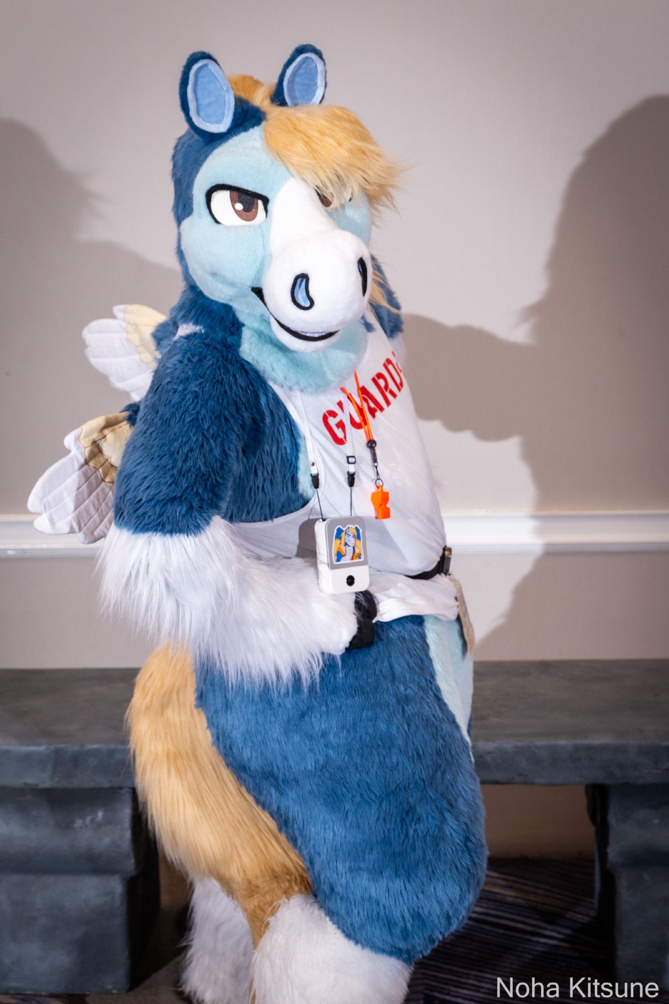 A photograph of a blue pegasus fursuiter at ConFuzzled 2024, standing in front of a grey bench with a plain white wall and cast shadows behind him. He's facing the camera and turned slightly to the side with hands on hips and wings visible.