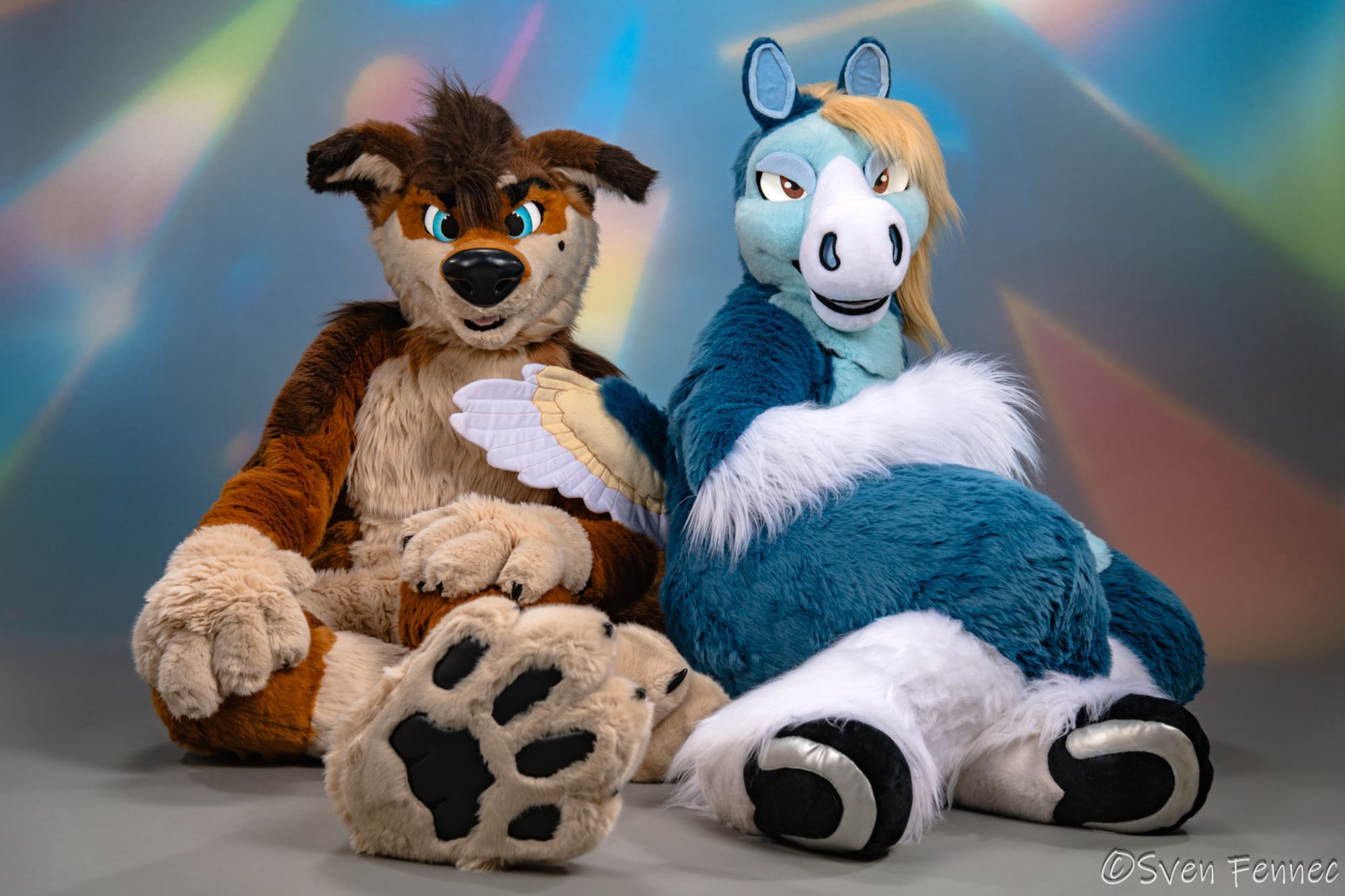 A studio photograph of two fursuiters sitting in front of an abstract 90s-style backdrop. On the left is a red wolf sitting with his footpaws out and his hands relaxed, and on the right is a blue pegasus sitting slightly turned with legs bent and both hooves visible.