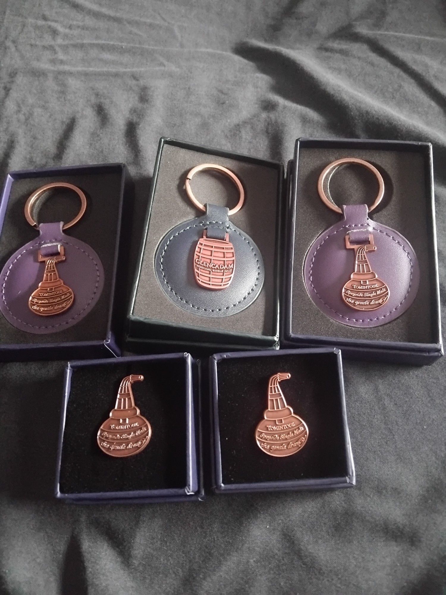 Photo showing three keyrings and two pin badges. Two of the key rings depict a pot still, and the other a barrel. The pin badges depict pot stills