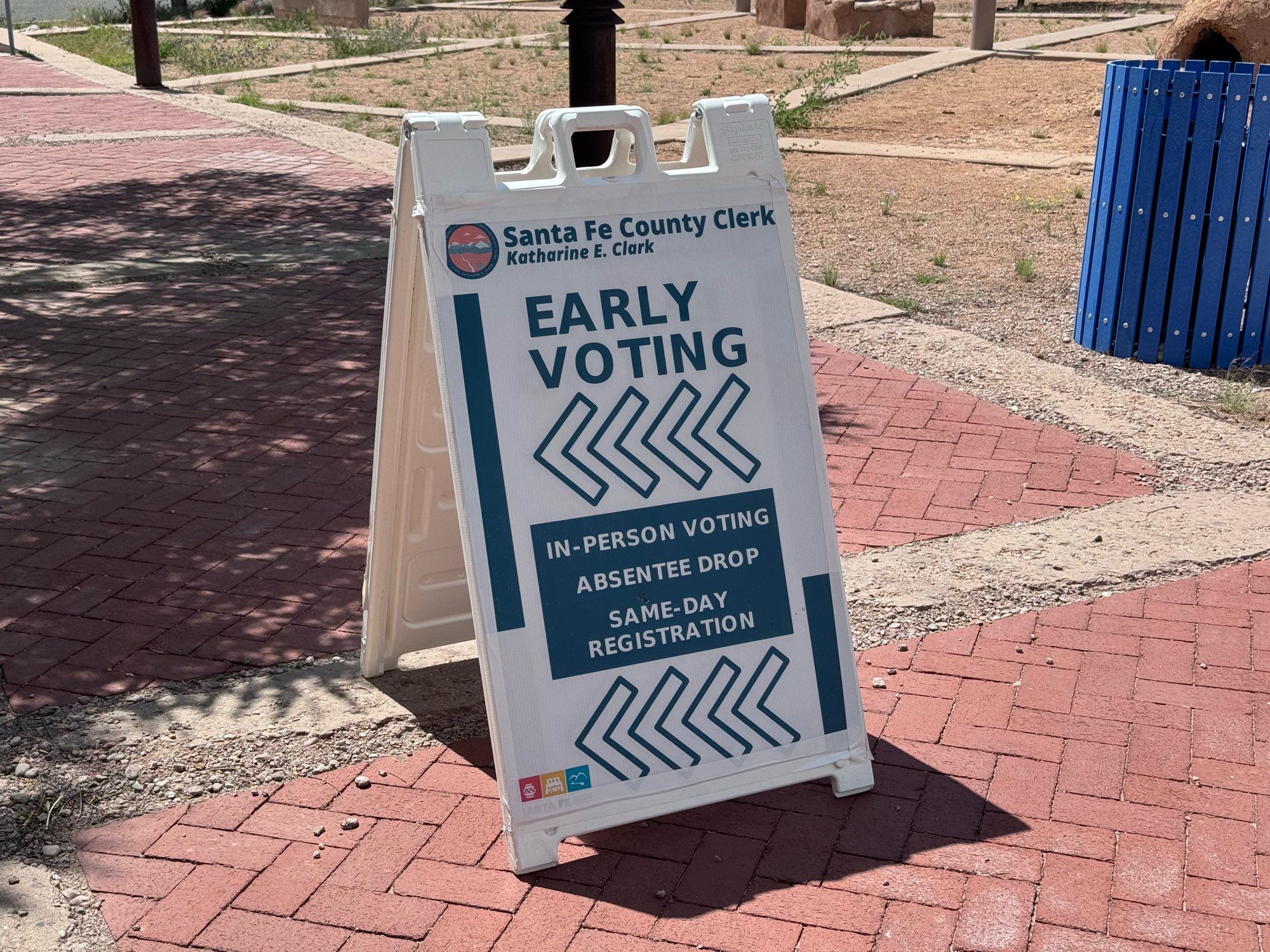 A folded sign set on a cement sidewalk that reads, in large letters “Early Voting”. Above, in smaller letters, is “Santa Fe County Clerk Katherine E. Clark”. On the bottom, in smaller letters, is “In-Person Voting”, “Absentee Drop”, and “Same-Day Registration”.