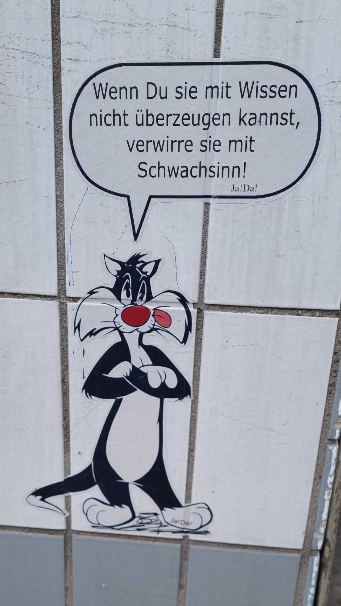 Pasteup auf einer gefliesten Hauswand. Comicfigur Silvester (der Kater). Sprechblase mit dem Text "Wenn Du sie mit Wissen nicht überzeugen kannst, verwirrse sie mit Schwachsinn!".