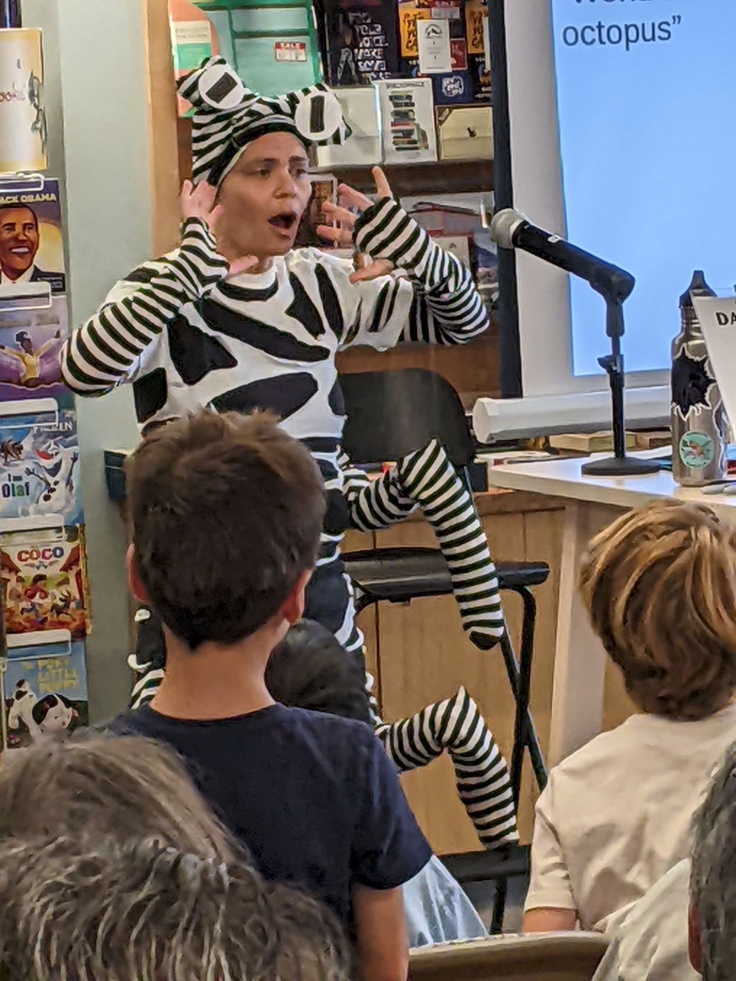 A closeup of Danna in her mimic octopus costume, face uncovered, in the midst of talking and gesturing with her hands. The back of some audience heads are visible in the foreground and the word "octopus" is visible on the projector screen next to her.