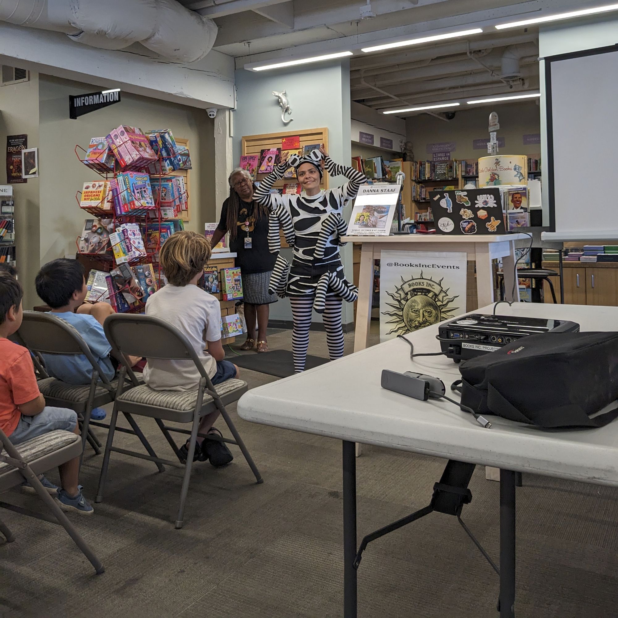 Danna begins her presentation in the bookstore, wearing her mimic octopus costume, but with the face covering rolled up to her forehead so her face is visible, smiling. A Books Inc bookseller stands behind her, and several children are visible from the back sitting in the audience.