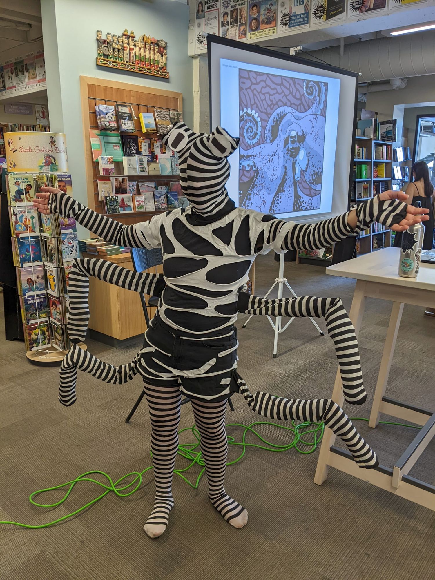 Danna Staaf stands in a bookstore in front of a projector screen with an octopus on it. She is dressed in a full-body mimic octopus costume: black and white striped tights on her legs, similarly striped stockings on her arms with finger holes cut out, and four additional stockings stuffed and pinned to her sides and connected to her arms with fishing line. Her face is also covered with a striped stocking with two protrusions on top of her head decorated to look like octopus eyes.