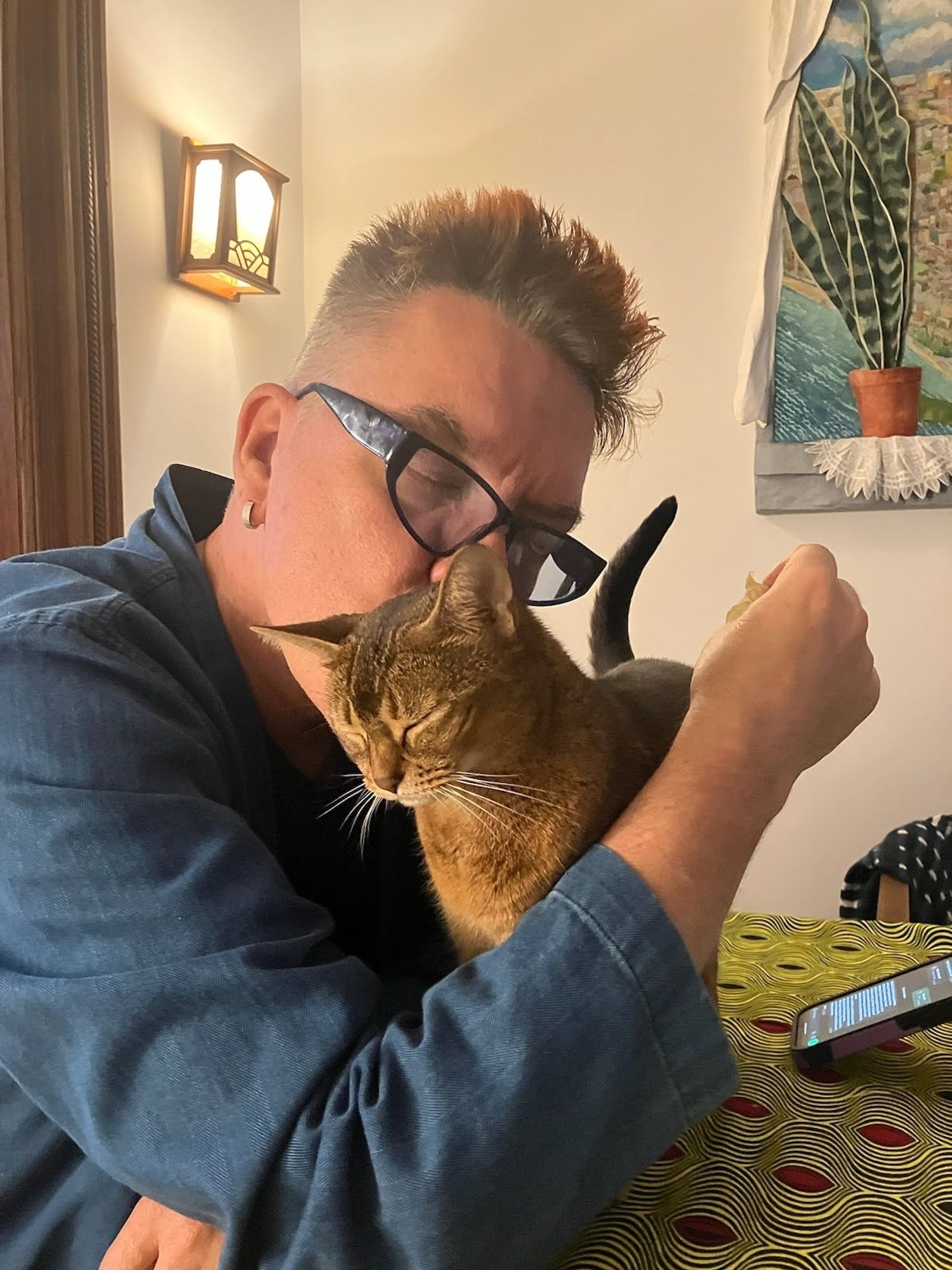 A middle-aged white man with spiky orange and gray hair and blue glasses embracing a Ruddy Abyssinian cat and kissing the back of her neck. Both man and cat have their eyes closed in a moment of shared tenderness.