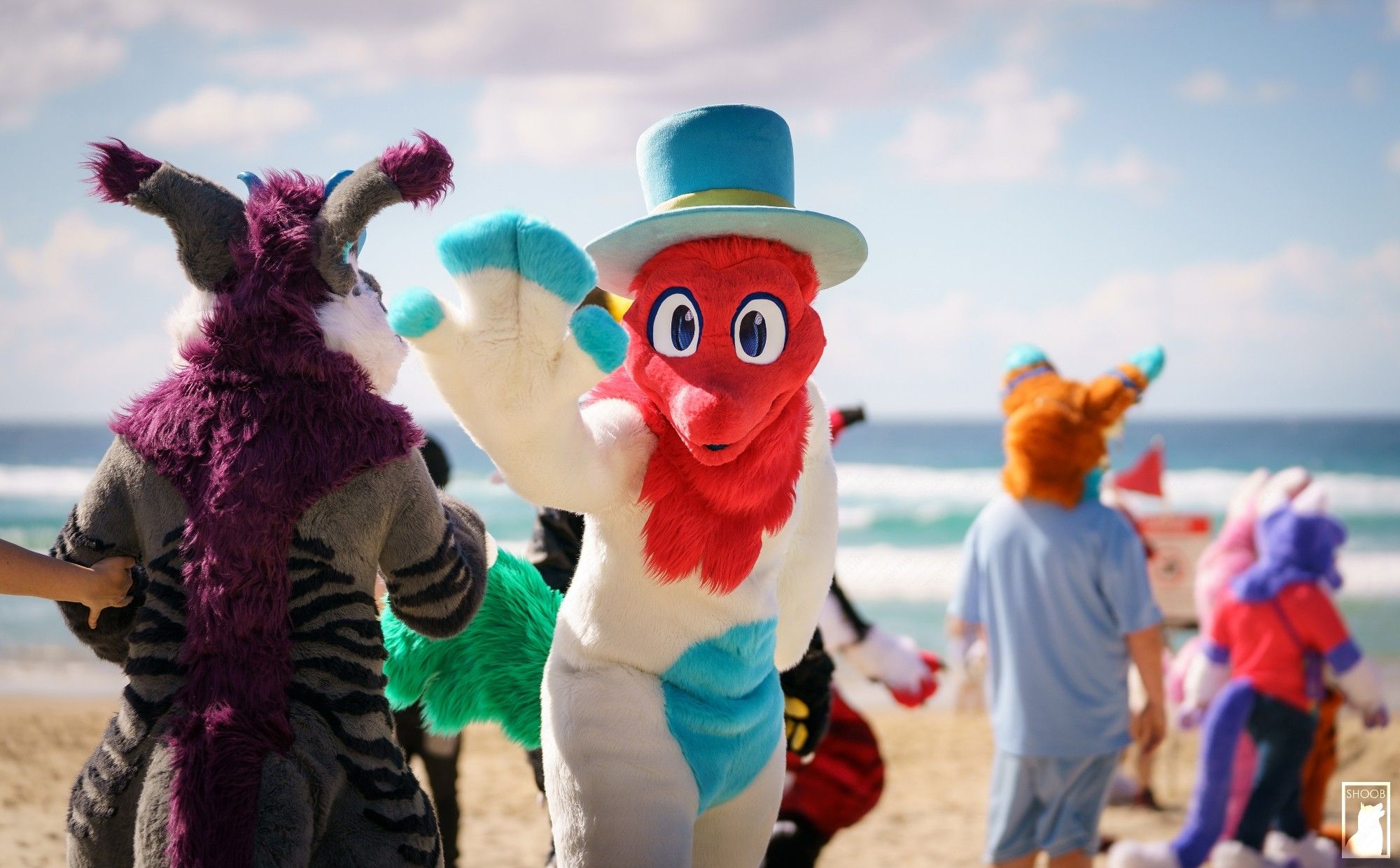 A photo of Dip waving at the camera during a Fursuit parade on the beach at FurDU, the Gold Coast Furry Convention