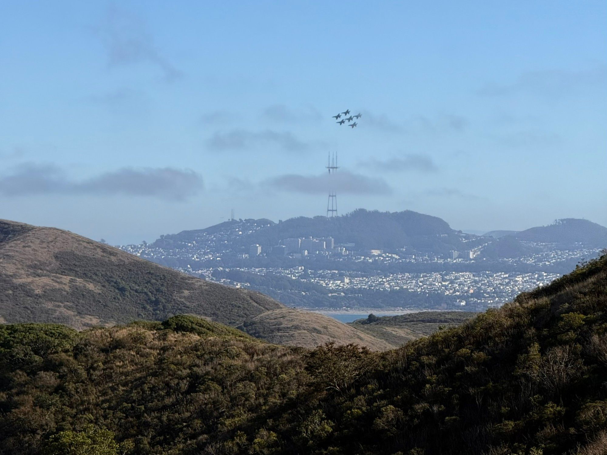 A couple hills in the foreground meet near the bottom middle of the image to form a wide shallow V. Riding from the valley is the San Francisco skyline from the north, crowned by Sutro Tower. The tower is partially obscured by a whiff of cloud. Five jets flying in tight formation are just above the tower. The sky is blue. Image courtesy of https://androiddev.social/@zachklipp 