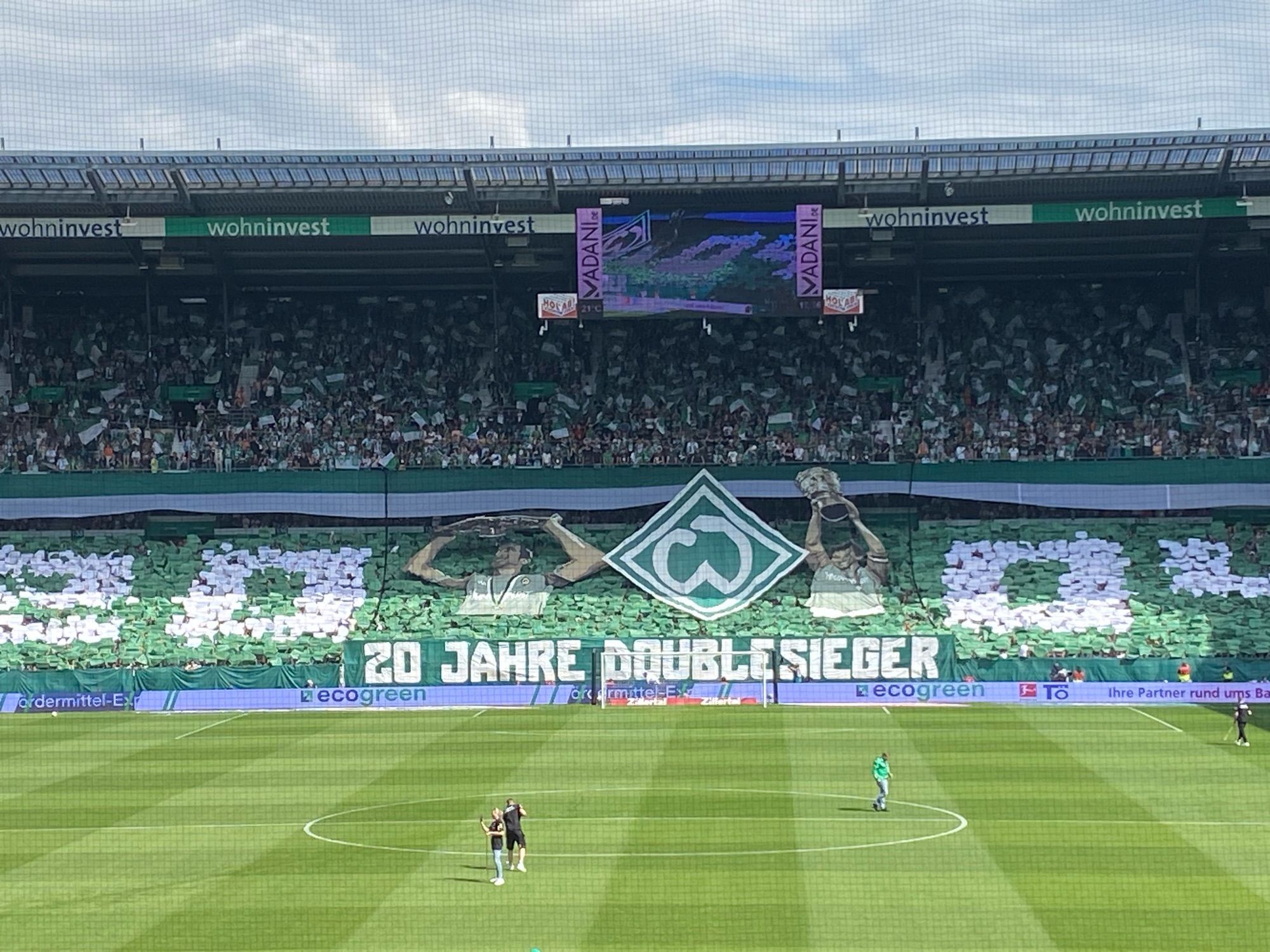 Die Ostkurve im Weserstadion frontal aufgenommen. Das untere Drittel zeigt den Rasen. Im Stehblock ist eine Choreo zu sehen. Eine weiße 2004 zwischen den Nullen ist links ein Werderspieler mit der Meisterschale, dann die Werderraute und rechts ein Werderspieler mit dem Pokal. 
Darüber der Oberrang mit Zuschauern, mittig ein Stadionmonitor, abschließend das Stadiondach und ein bewölkter Himmel