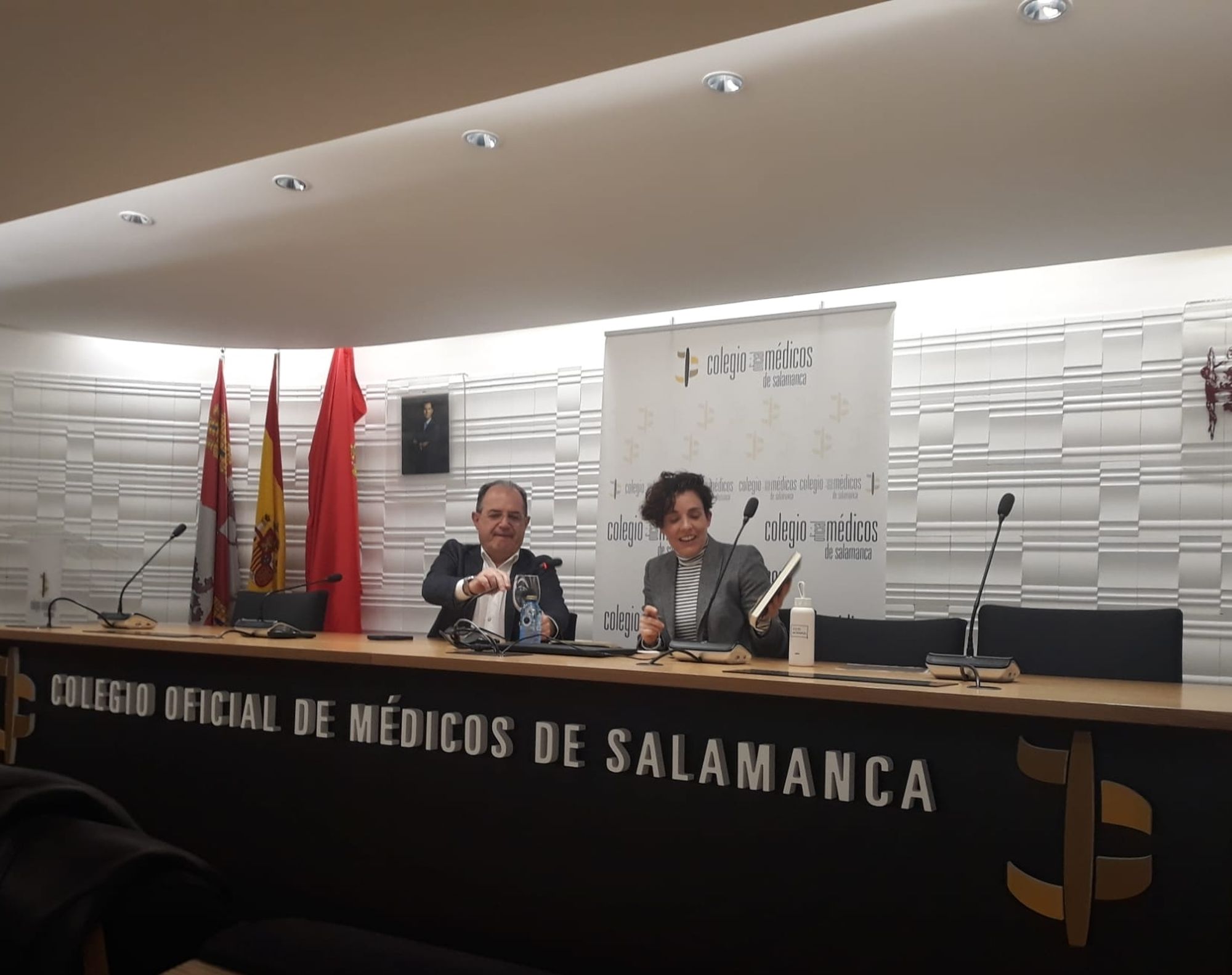 Fotografía de la charla. Presenta el presidente del Colegio. Detrás se ve un cartel y unas banderas institucionales. Yo sostengo un libro.