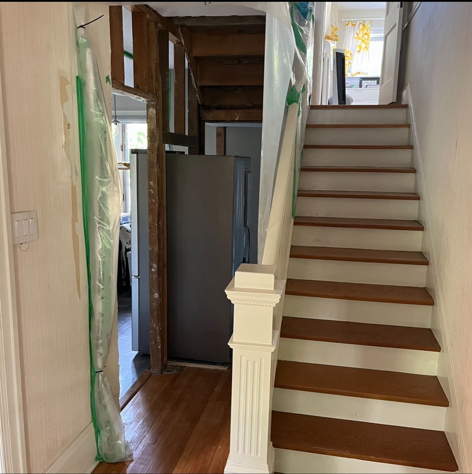 On the right, a beautiful old stair case with white trim, wood treads, and a classic newel post. On the left, the walls and ceiling are stripped away, revealing old 2x4 framing and the underside of the stairs. There’s a refrigerator right in the middle of everything, blocking the access to the back door.