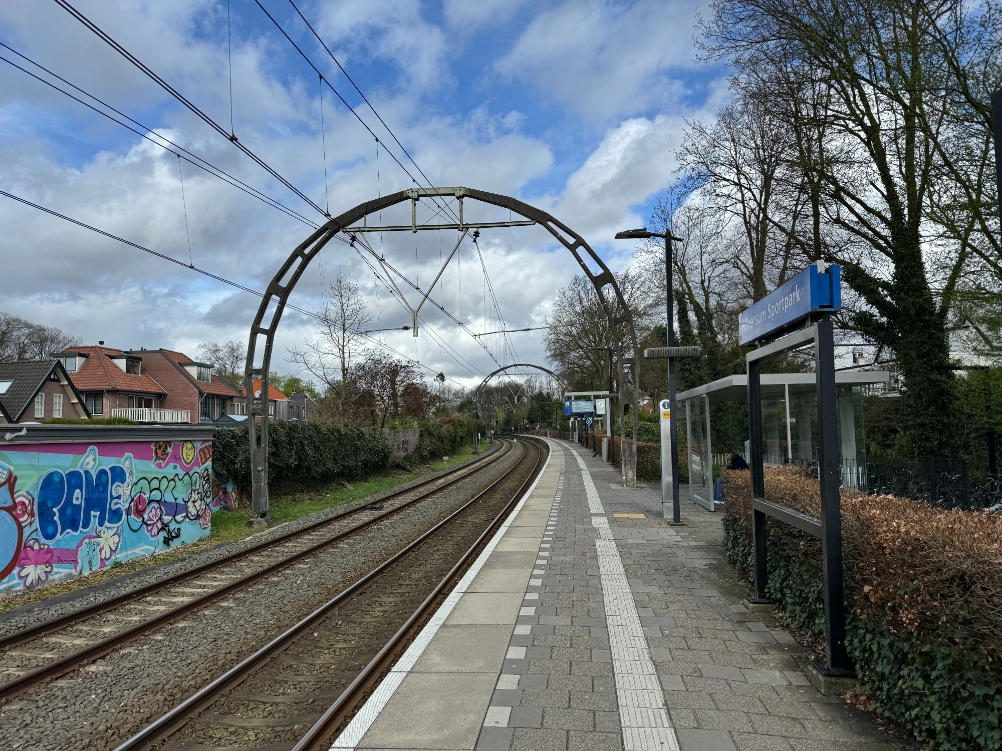 Photograph of arch-shaped supporting pole for overhead catenary wires over two railroad tracks at a passenger rail station near Utrecht, Netherlands. In the background: single story suburban houses, a graffiti-covered low wall, trees and hedges, blue sky with some fluffy clouds.