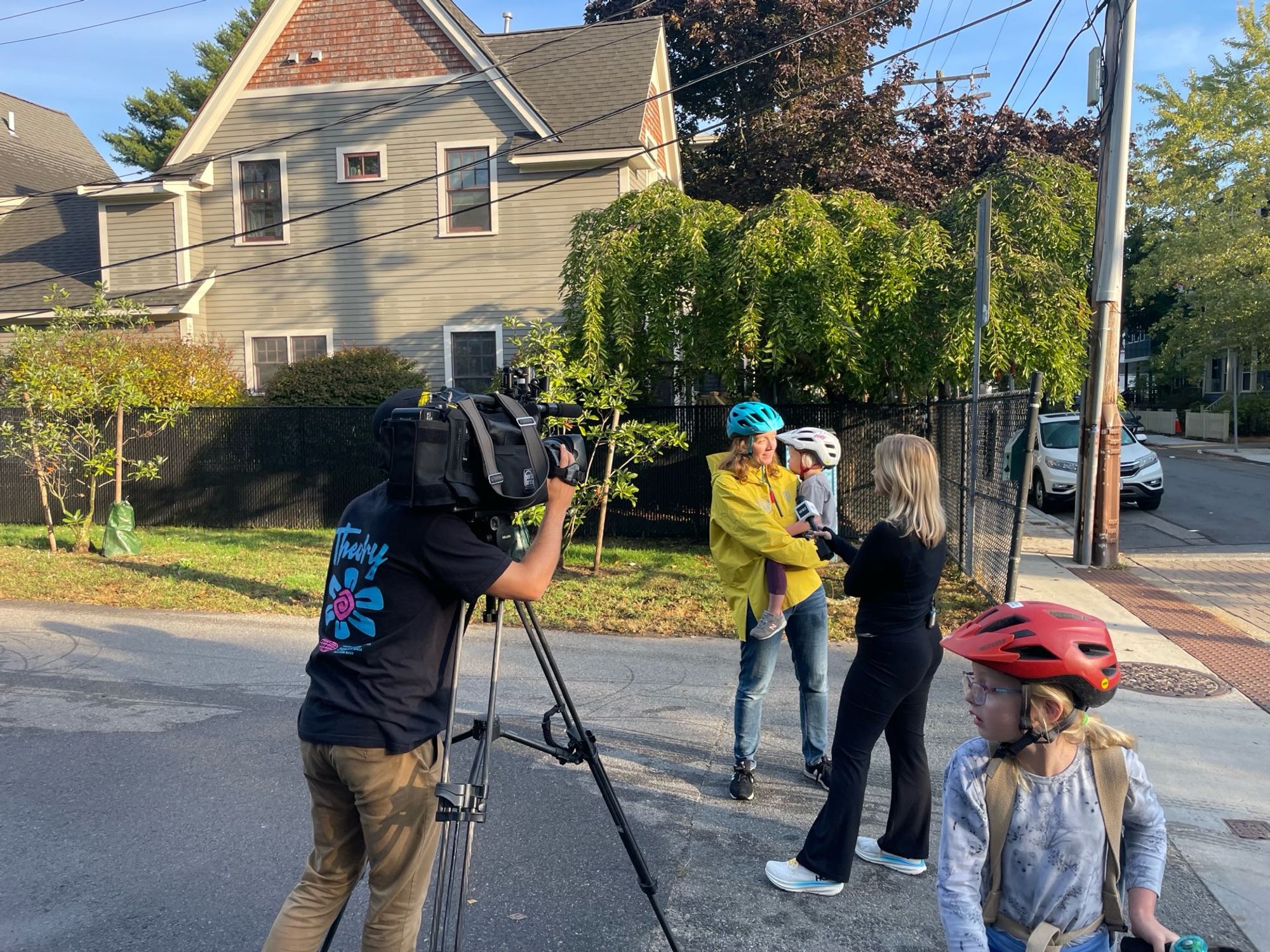 Woman with bike helmet holding a kid, being interviewed on camera by a reporter 