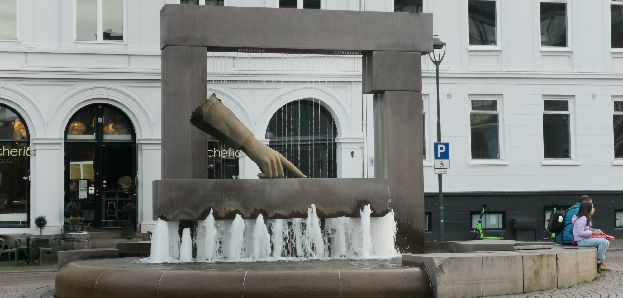 Ein kleiner Springbrunnen, darüber in einem Viereck aus Metall eine Hand ebenfalls aus Metall. Der Zeigefinger zeigt Richtung Wasser